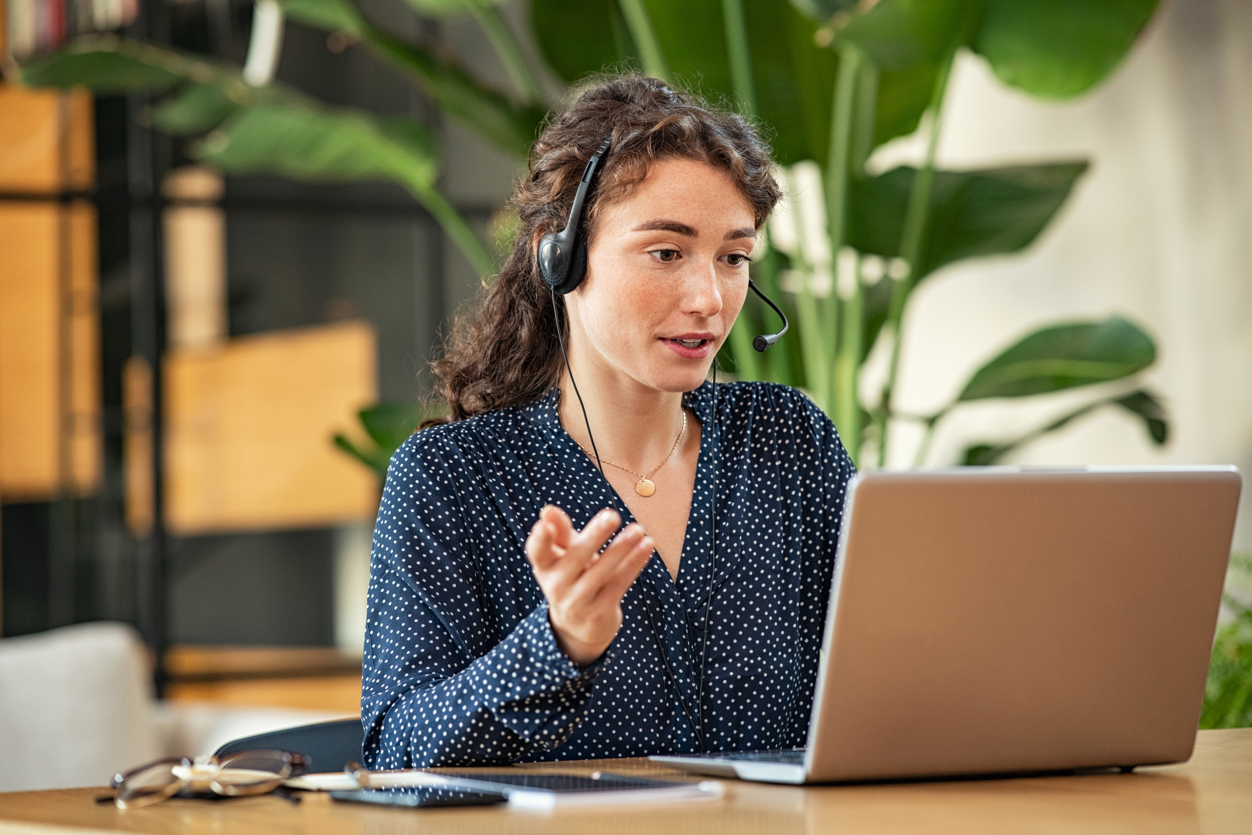 A woman wearing a navy blue polka dot shirt and a headset with a microphone, sitting at a wooden desk with a laptop, in a bright office environment with large green plants.