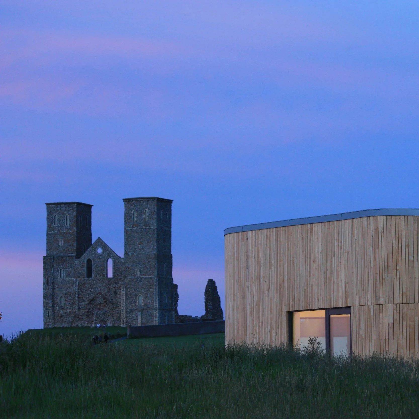 A historic stone church with two towers and a modern wooden building with a curved roof, set against a dusky sky with pink and purple hues, and a grassy foreground.
