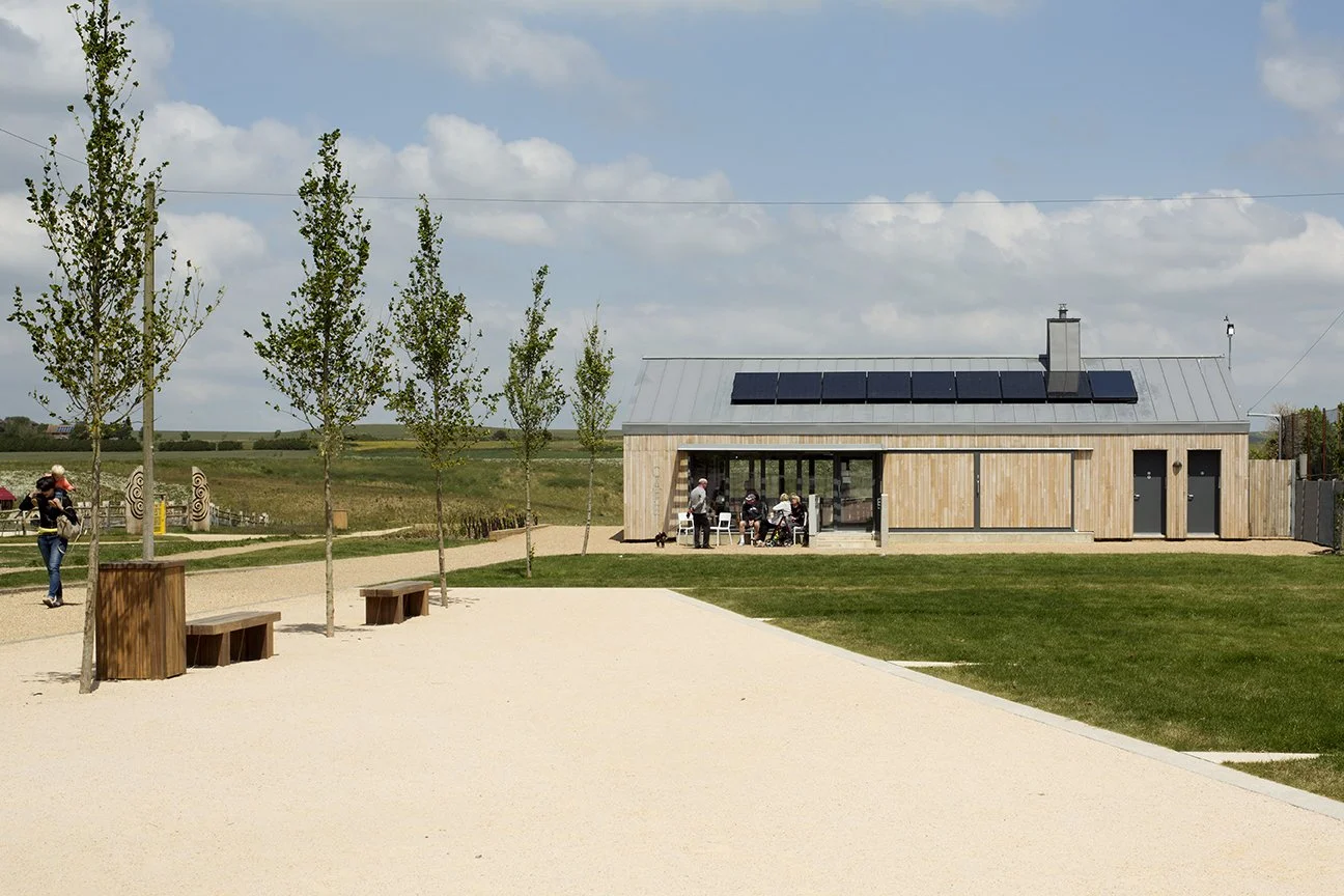 A modern, small building with solar panels on its metal roof, set in a park with walking paths, benches, and green grassy areas. There are people sitting outside and a person walking with a dog.