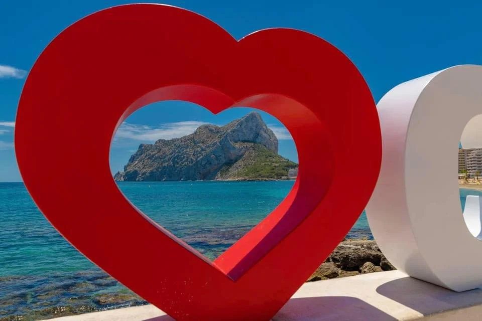 Large red and white sculpture spelling 'LOVE' with a heart shape in the middle, overlooking a beach with turquoise water and a mountainous island in the background.