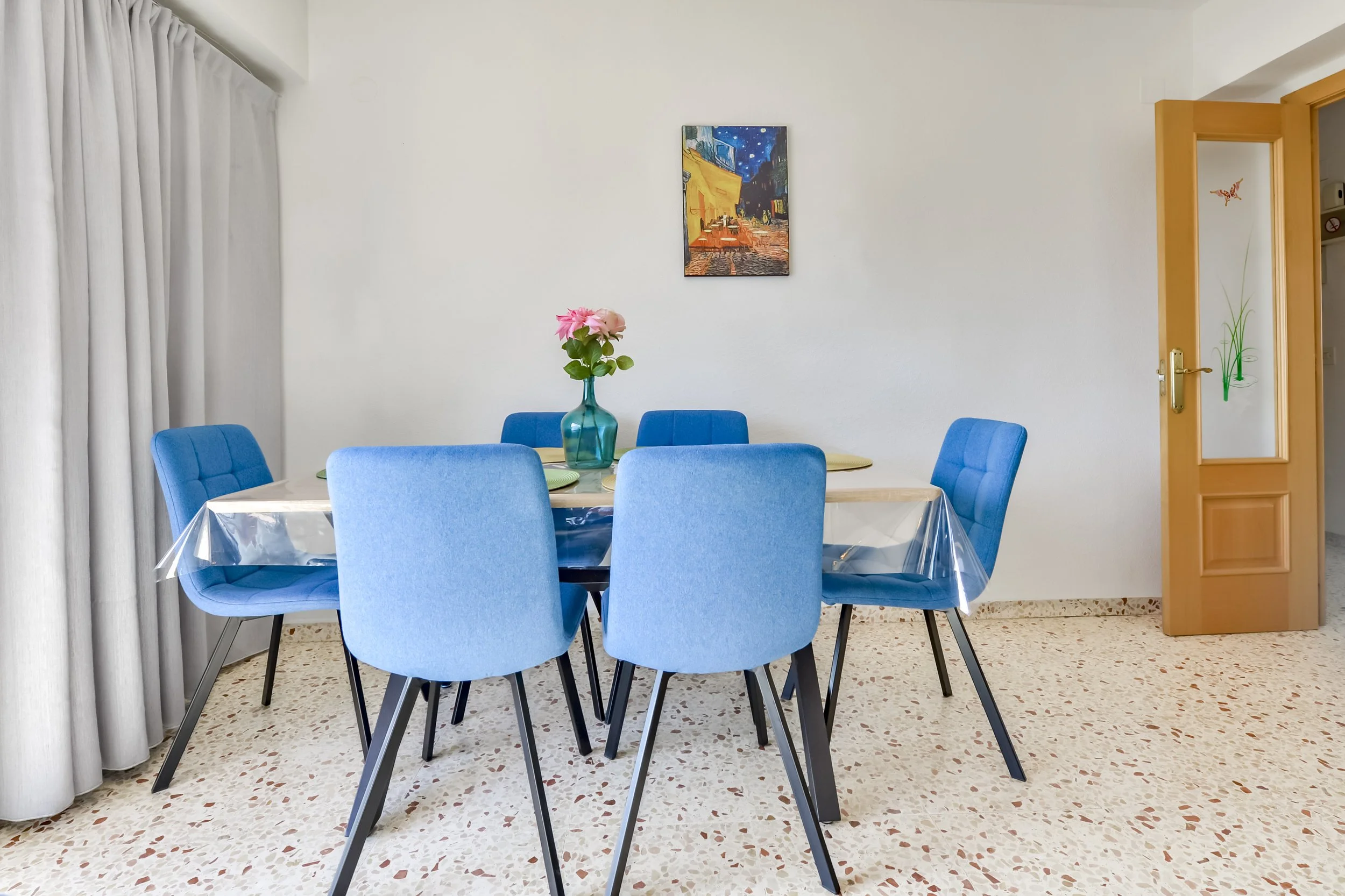 Eethoek van het vakantieappartement in Calpe met blauwe stoelen, roze bloemen en kleurrijk wandkunstwerk.
