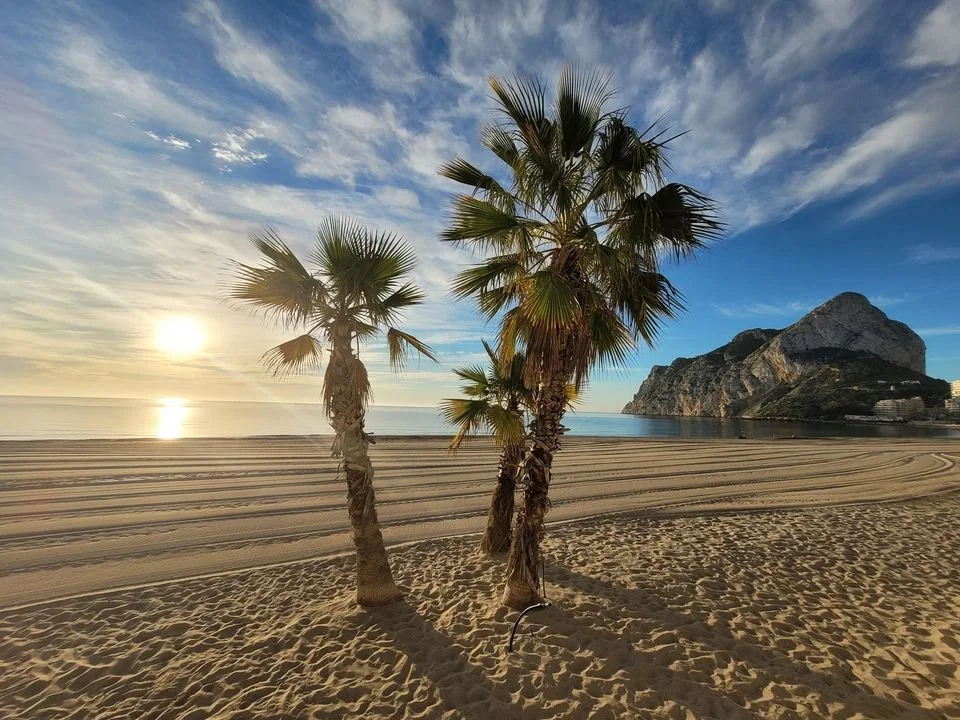 Zonsondergang boven het zandstrand van Calpe, met twee palmbomen op de voorgrond, de rustige Middellandse Zee en het silhouet van het eiland achteraan. Een idyllisch uitzicht dat je elke avond beleeft vanuit ons vakantieappartement bij Belgische eige