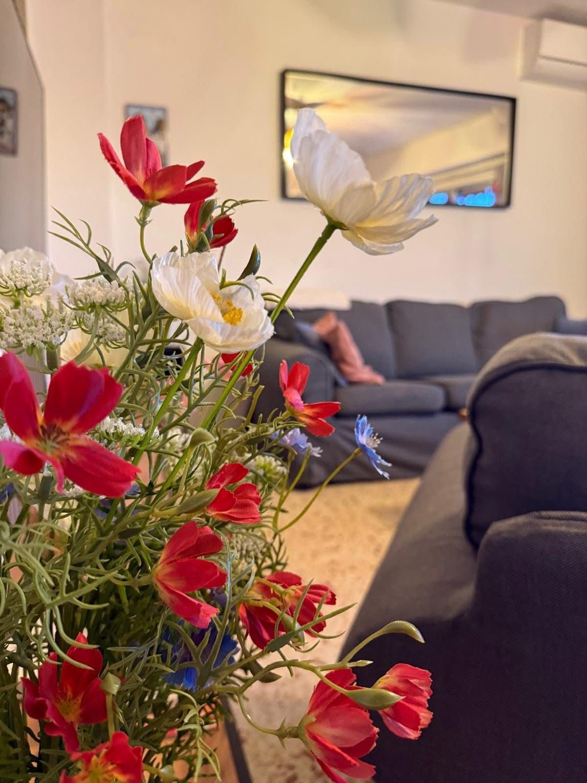 Close-up of a colorful flower bouquet with white, red, and blue flowers in a living room with a dark gray couch and a wall mirror.