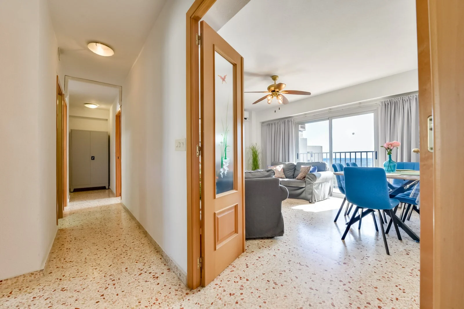 Inkomhal en leefruimte van het vakantieappartement in Calpe met houten deur, terrazzo vloer, grijze sofa en ronde eettafel met blauwe stoelen.