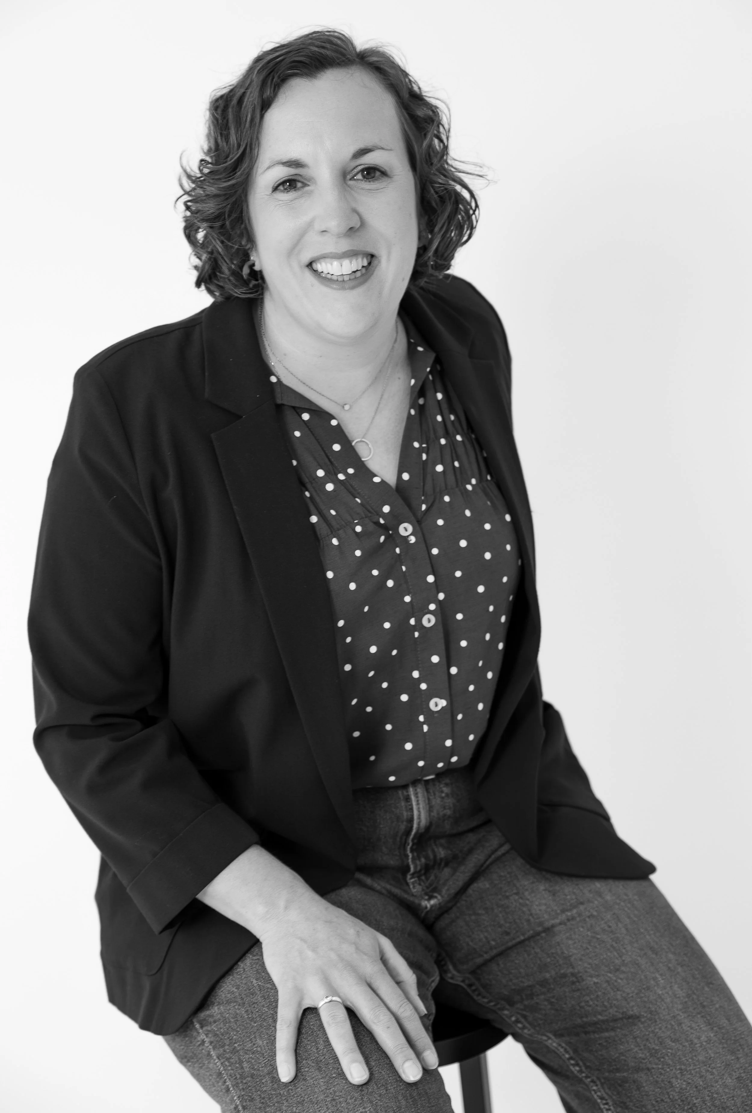 Black and white portrait of a woman with curly hair, wearing a blazer, polka dot shirt, and jeans, sitting with one hand on her knee, smiling and looking at the camera.