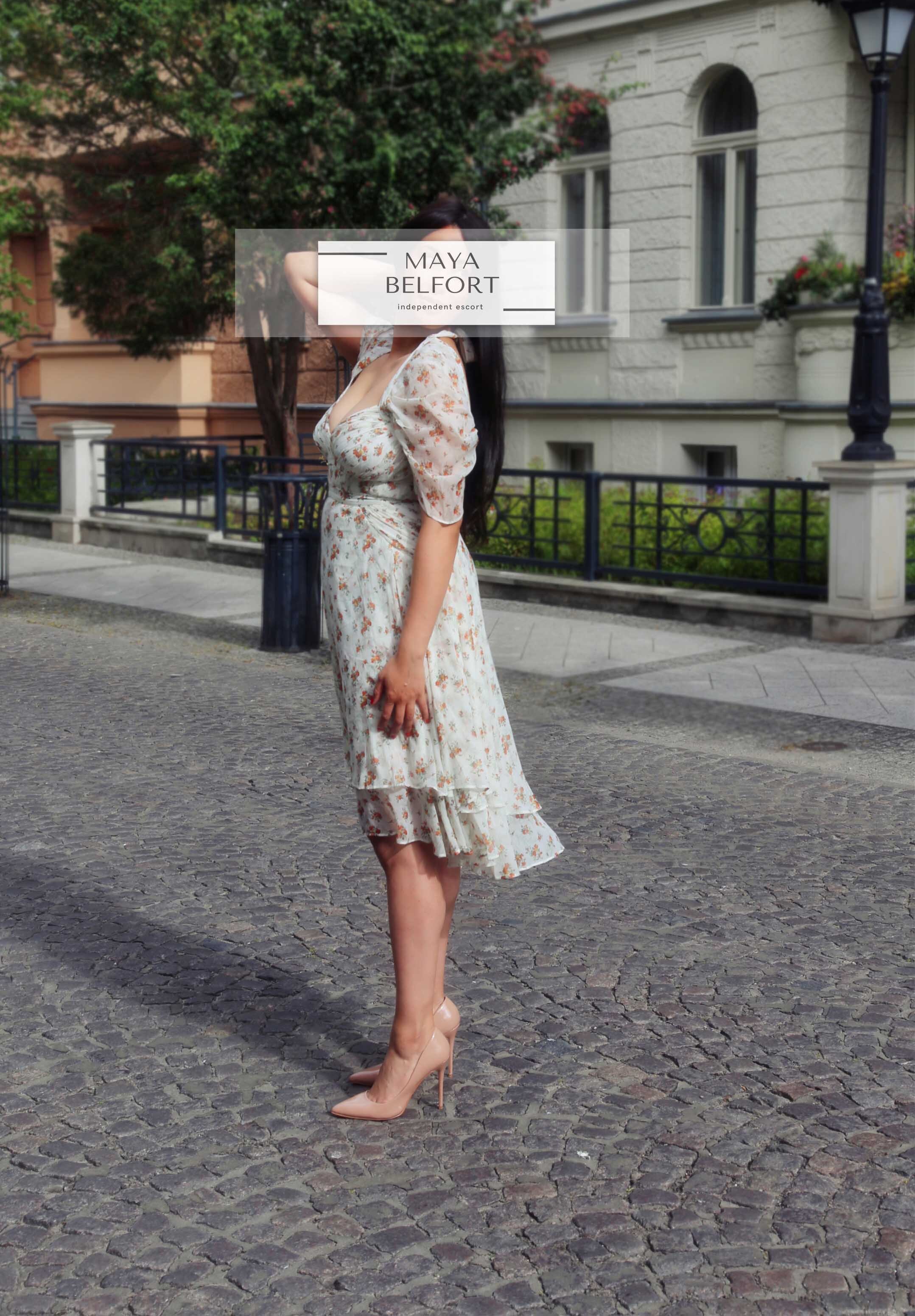 maya Belfort your escort europe stands on a pavement in a metropolis wearing a floral dress and bright pumps