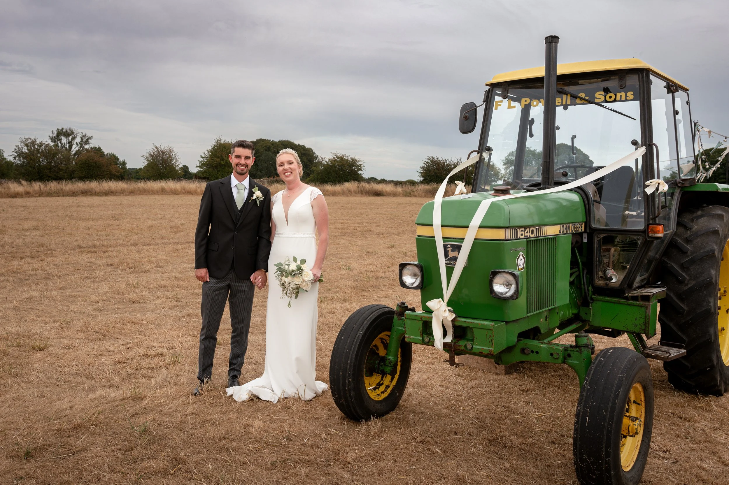 A newlywed couple in wedding attire standing outdoors next to a green John Deere tractor decorated with white ribbons.