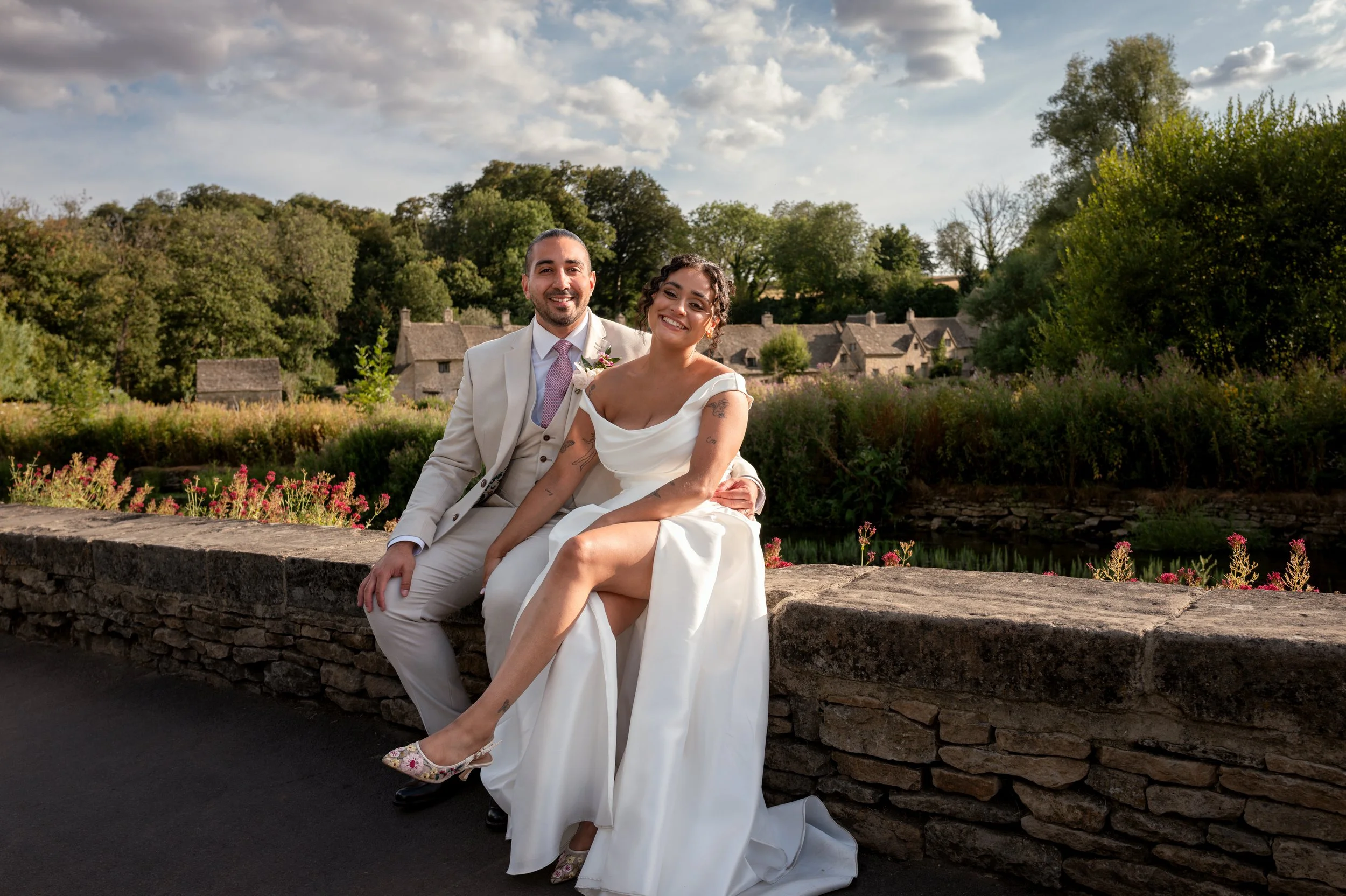 A newly married couple sitting on a stone wall in a garden, smiling at the camera, with trees and a village in the background on a partly cloudy day.