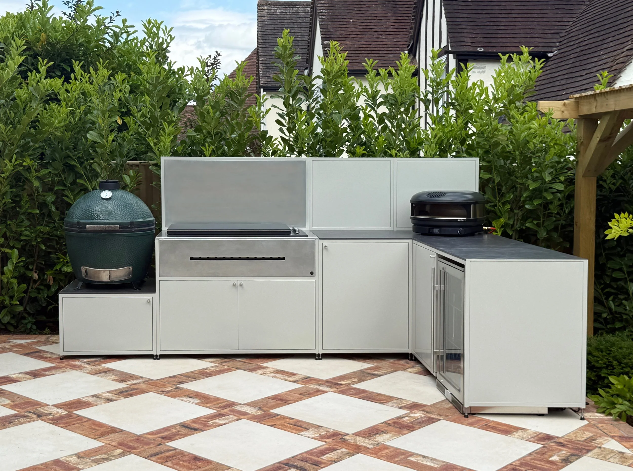 An outdoor kitchen with a white countertop, a green egg-style grill, a black Kamado-style grill, and a microwave oven set on a tiled patio with a checkered pattern. Green bushes and a house with a pitched roof in the background.