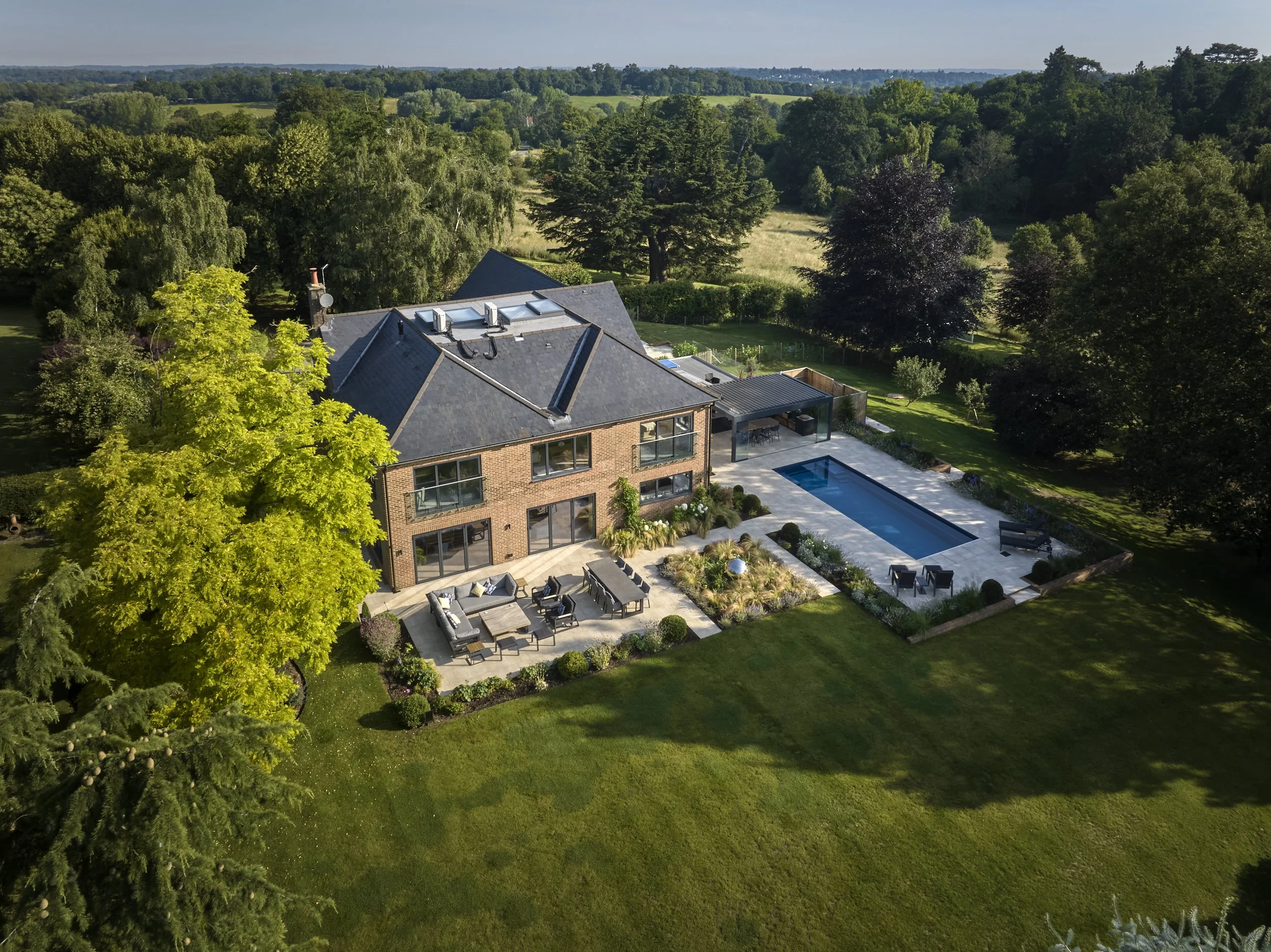 Aerial view of a modern house with a backyard that includes a pool, outdoor seating, and landscaped garden, surrounded by lush green trees and open fields in the distance.