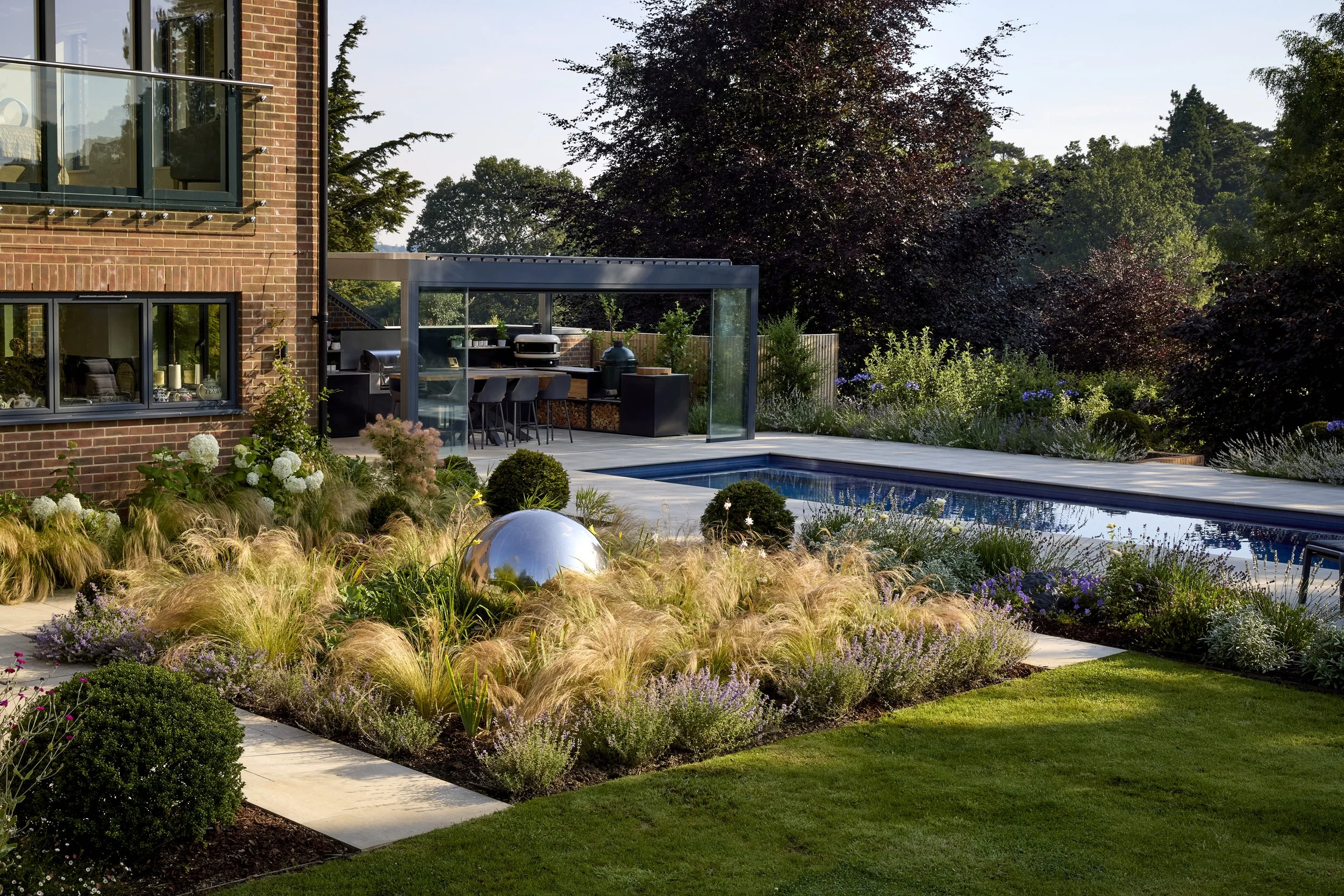 A modern house with a brick facade, large windows, and a glass-enclosed outdoor kitchen area. The backyard features a swimming pool surrounded by various plants, flowers, and shrubs, with a lawn and a patio area.