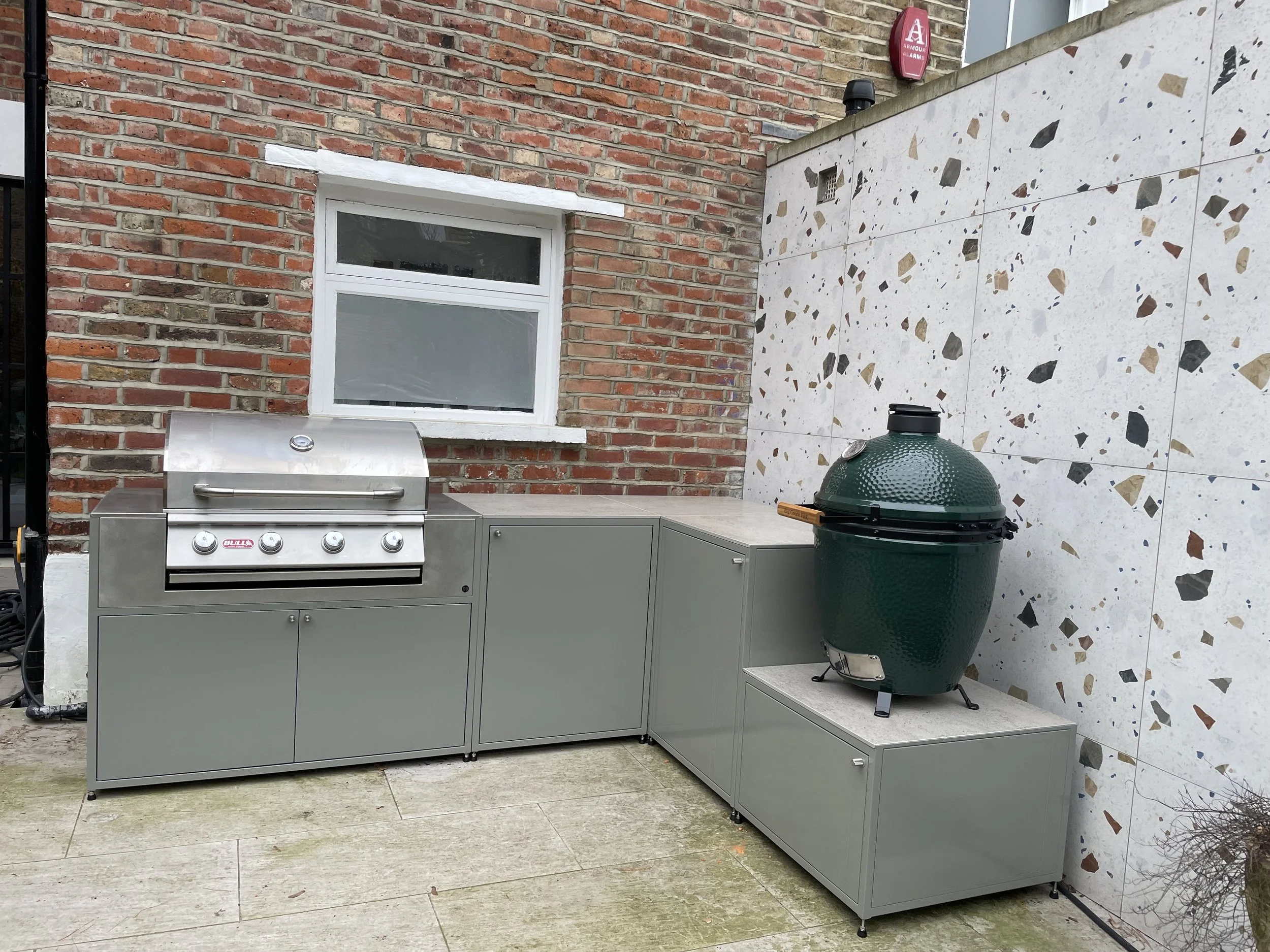 A backyard patio with a red brick wall, a white window, a stainless steel grill, a green ceramic smoker, gray cabinetry, and a terrazzo tiled wall.