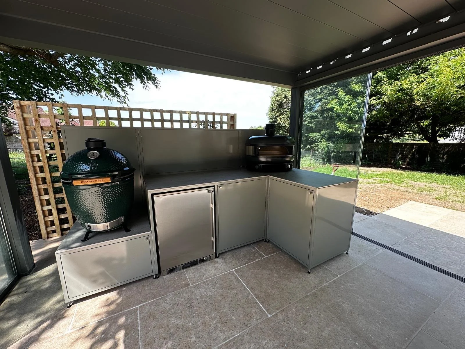 Modern outdoor kitchen with a green ceramic grill, a black pizza oven, gray storage cabinets, and a view of a backyard with grass and trees.