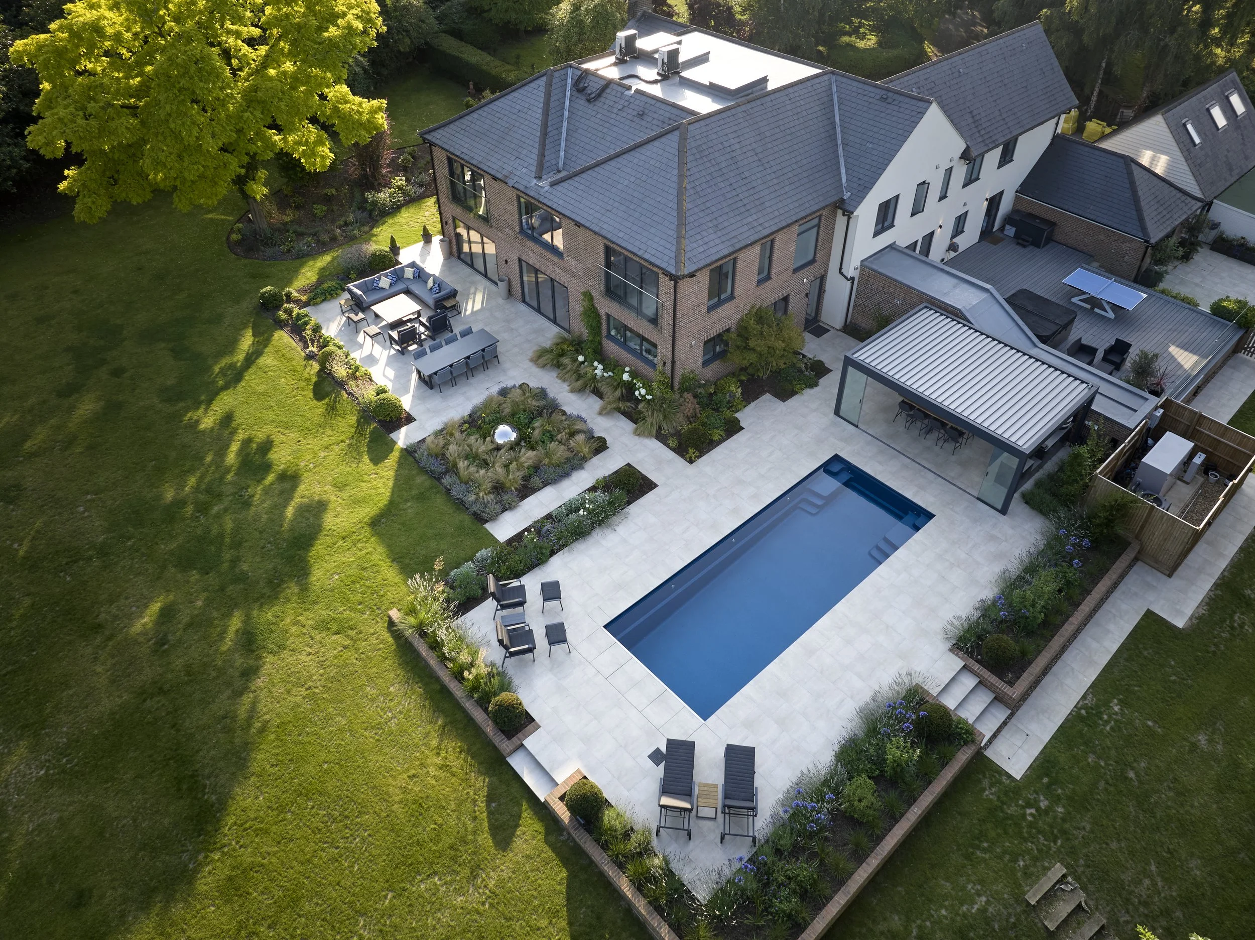 Aerial view of a modern house with a backyard patio, a pool, outdoor kitchen, canopy and outdoor seating area surrounded by a green lawn and trees.