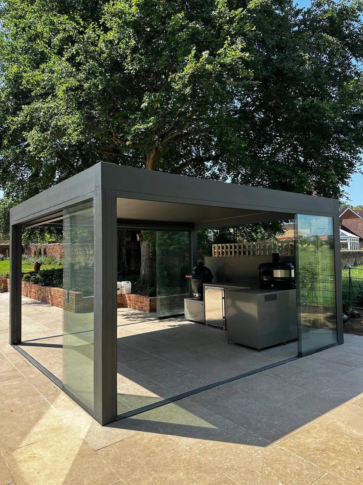 A modern outdoor barbecue shelter with glass walls and a flat roof, situated on a patio with a large tree and garden in the background.