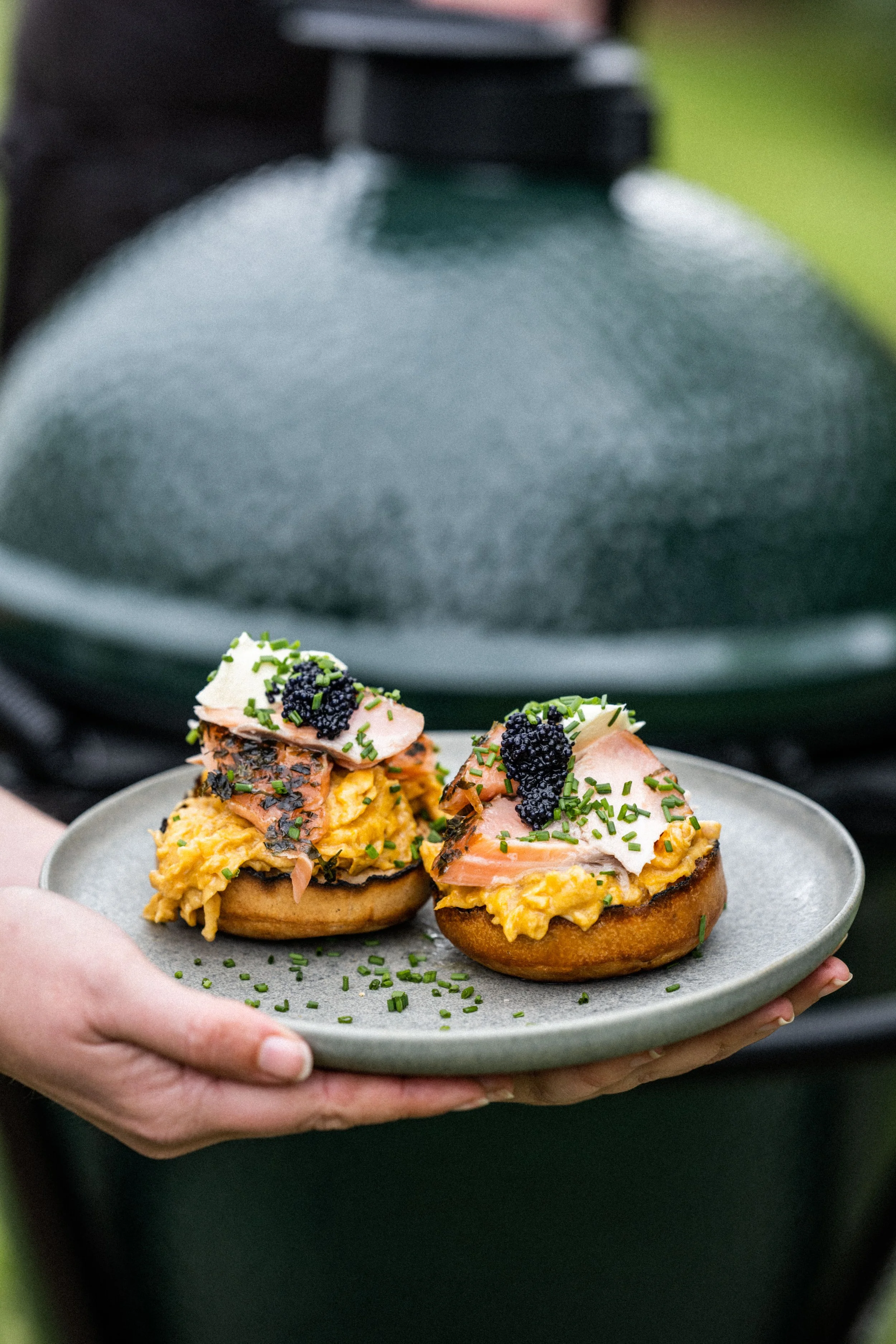 Person holding a plate with two gourmet breakfast toasts topped with scrambled eggs, smoked salmon, chives, and caviar.