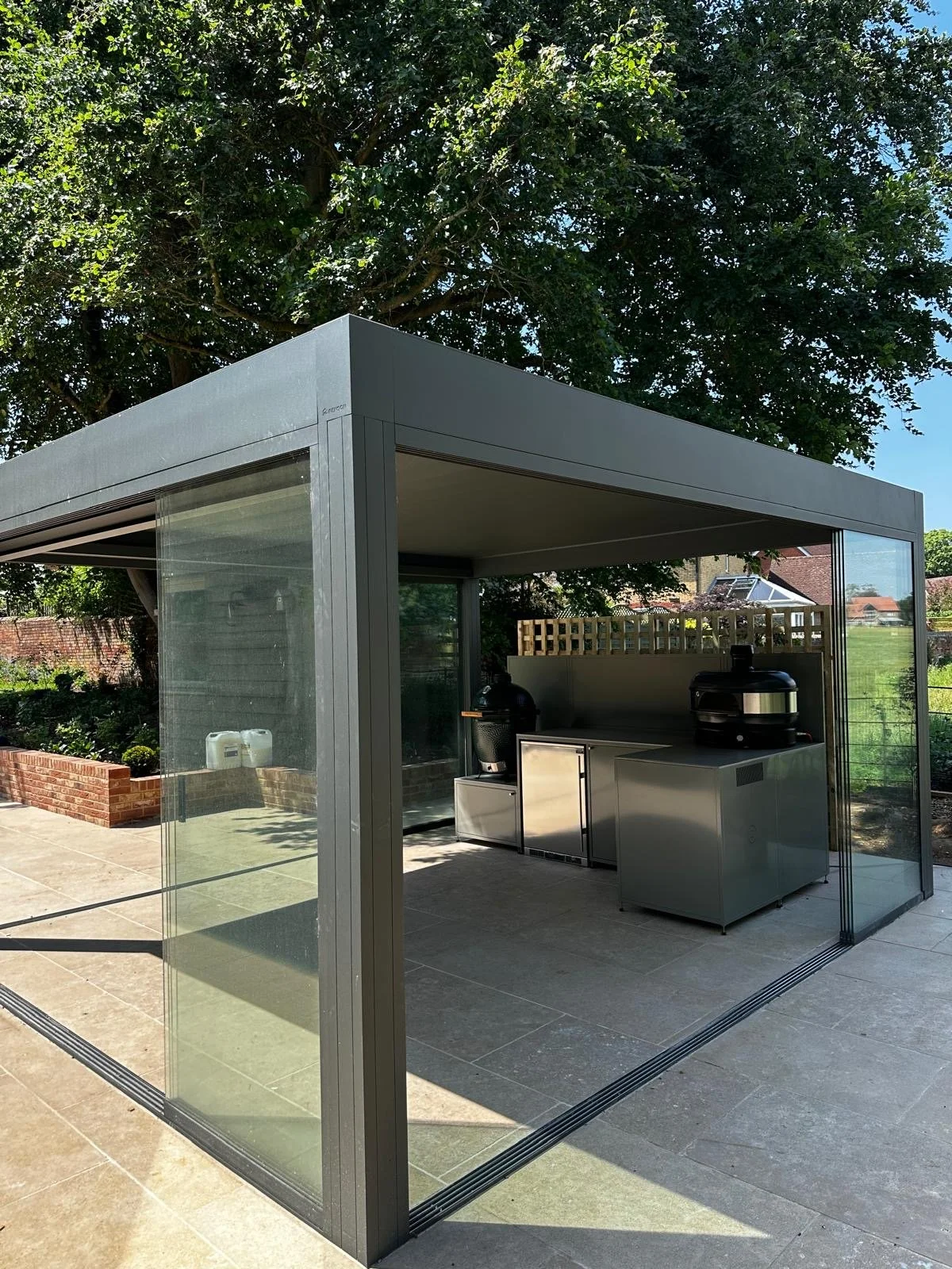 A modern outdoor glass and metal patio structure with a grill and outdoor kitchen area, surrounded by a garden and trees.