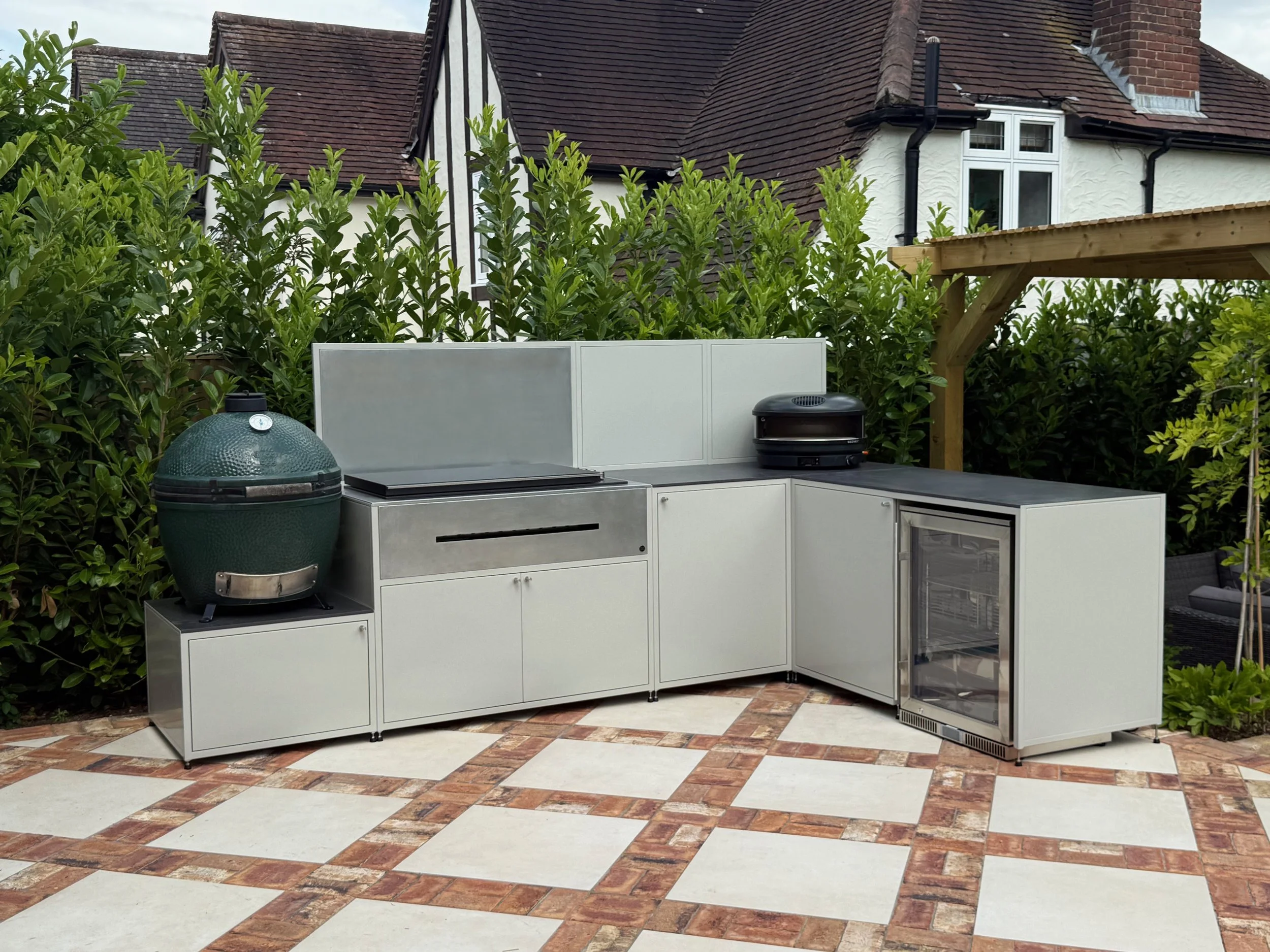 An outdoor patio with checkered red and white brick flooring and a built-in outdoor kitchen with a green ceramic grill, white cabinets, and a black countertop. There are lush green bushes behind the kitchen and a house with steeply pitched roof and b