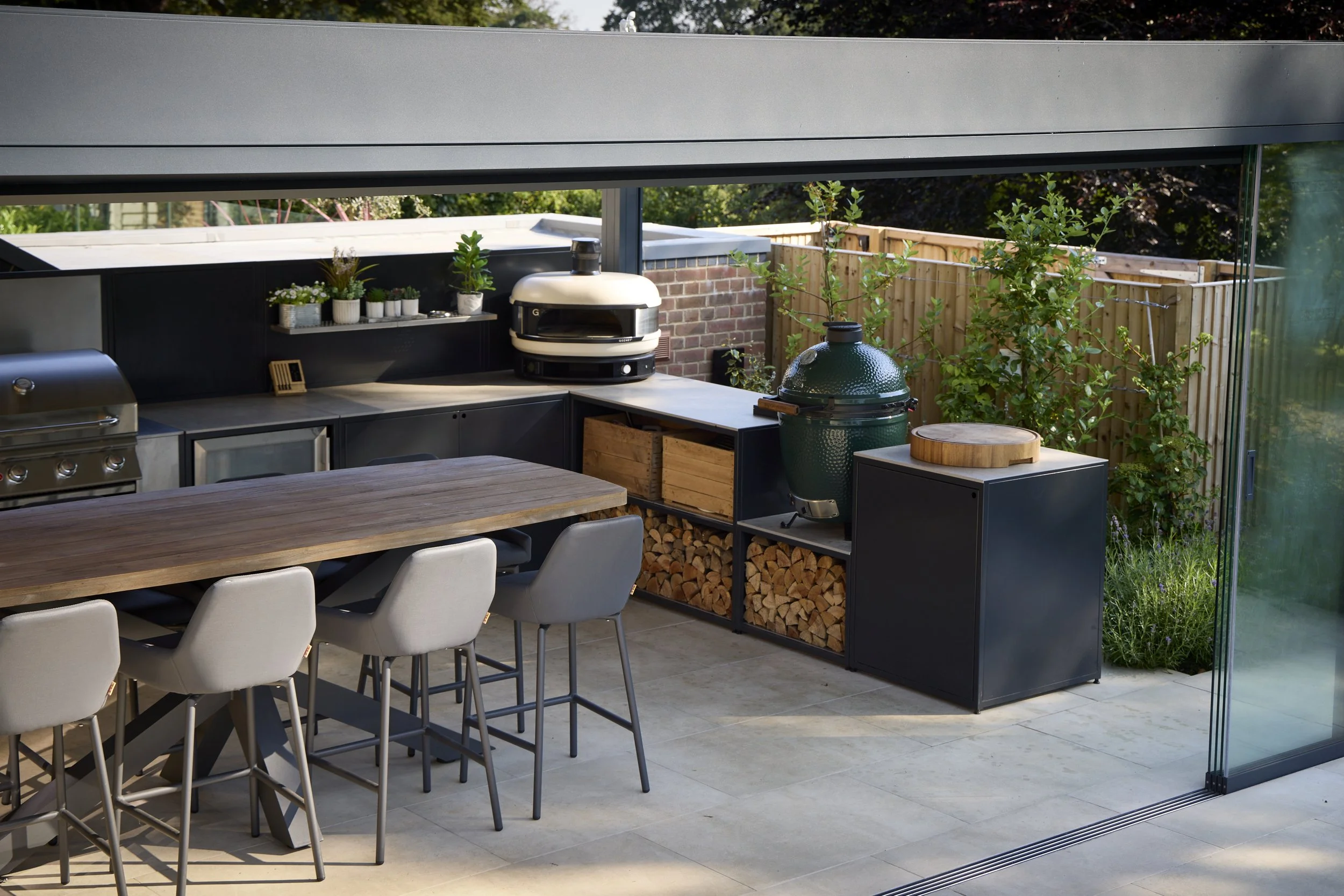 Modern outdoor kitchen under a weatherproof pergola with a wooden dining table, gray chairs, and built-in black cabinets containing a grill, a wood fire oven, and firewood storage, surrounded by a wooden fence and lush green trees.