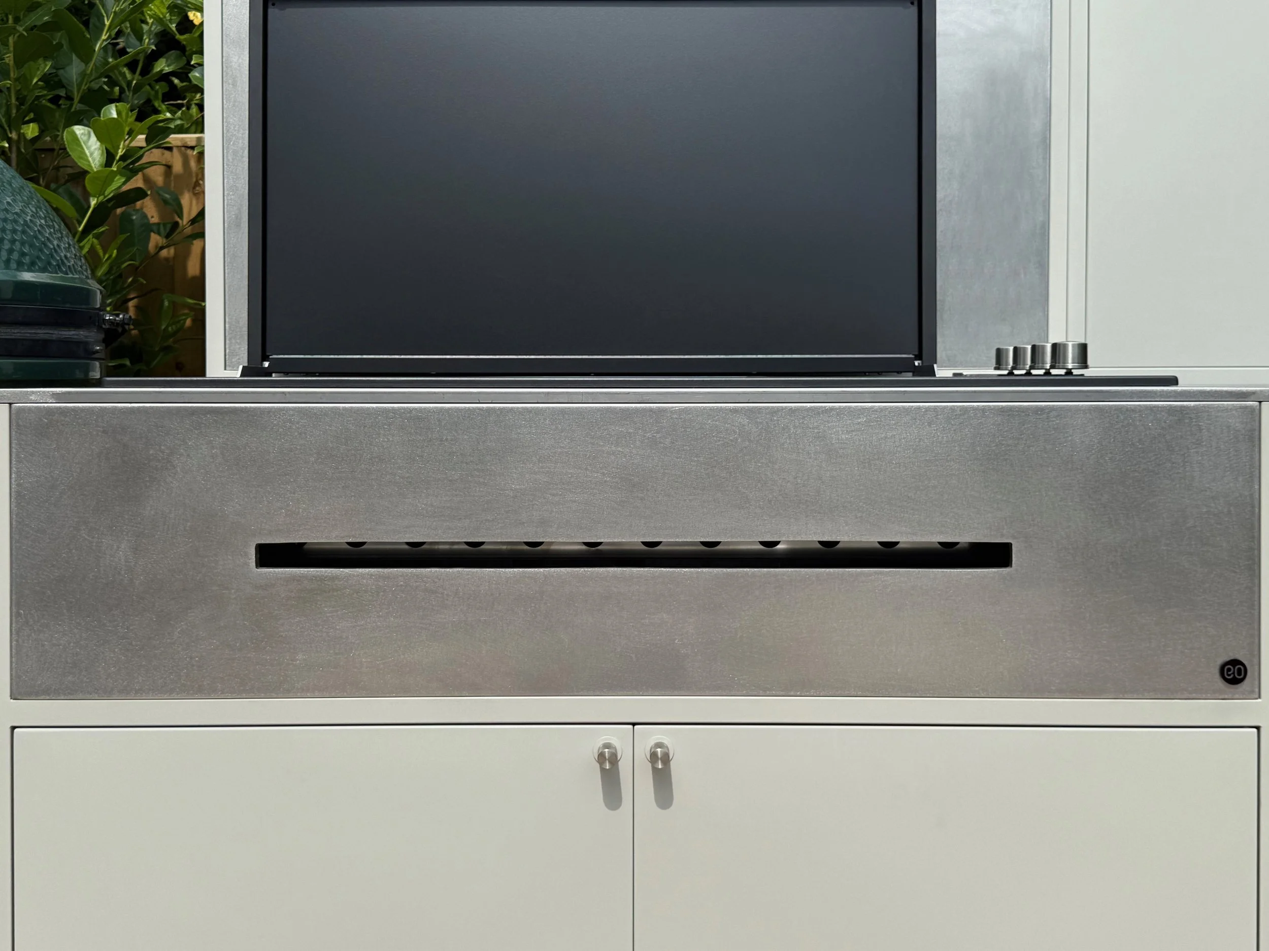 A modern outdoor kitchen grill with stainless steel doors, four control knobs, and a black cover hanging over the grilling area. There are green plants and a wooden fence in the background.