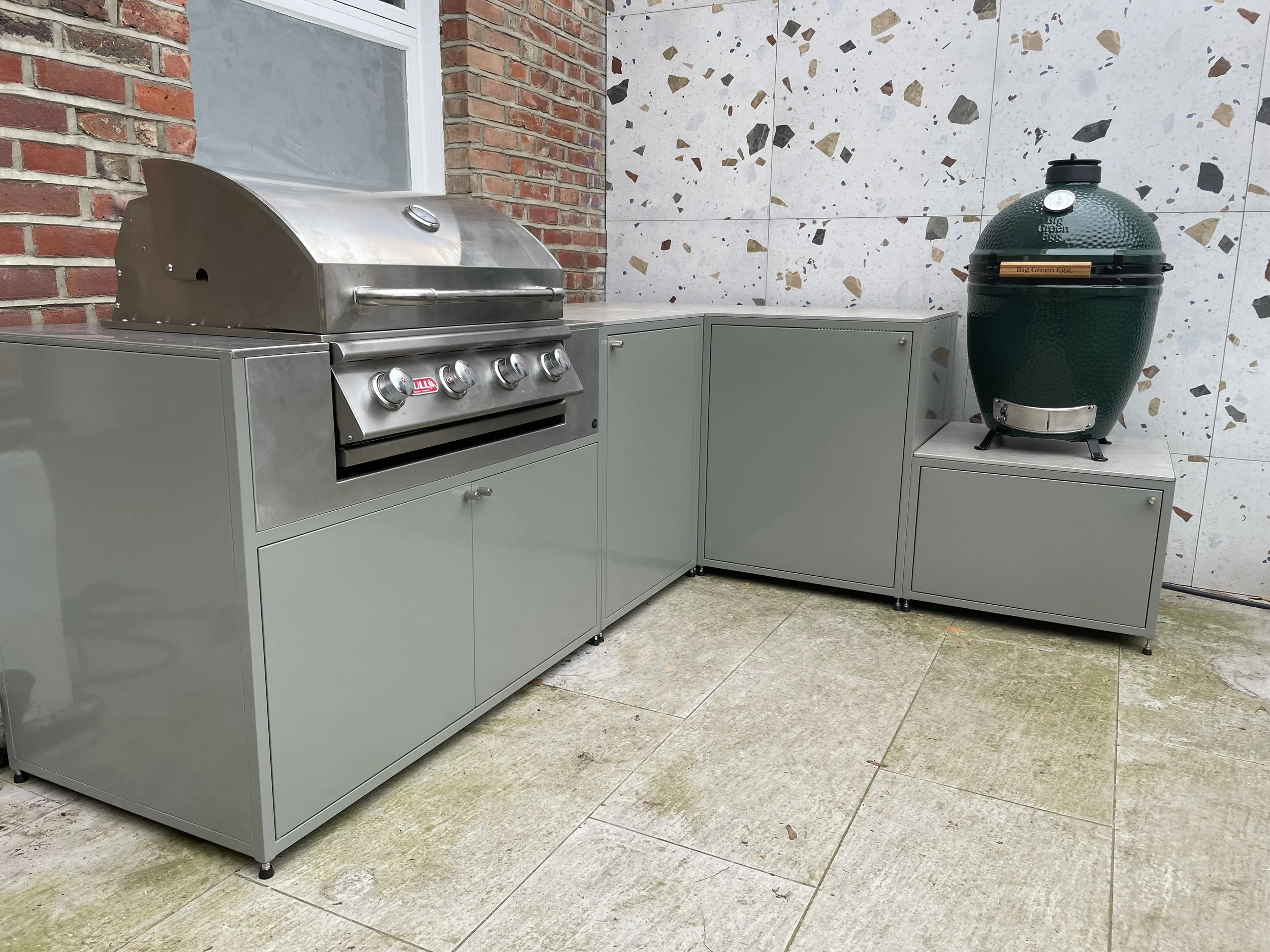 An outdoor kitchen area featuring a stainless steel gas grill with control knobs and a large green ceramic egg-shaped smoker on a table, against a background of a brick wall and terrazzo patterned wall tiles.