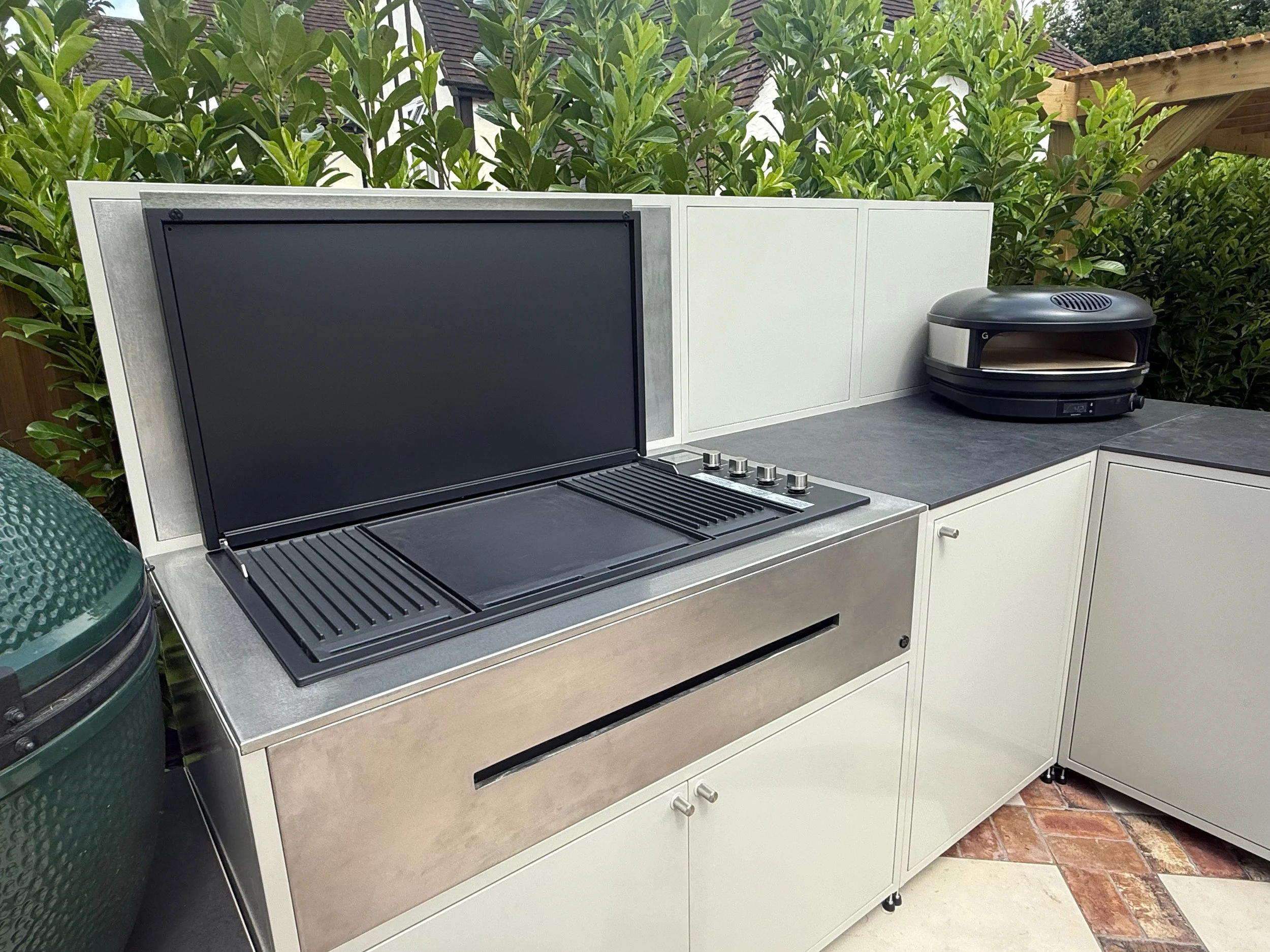 An outdoor kitchen area with a silver grill, a black grill lid, and a white countertop with white cabinets below. There is an oven or stove with dials, a black appliance on the countertop, a large green water storage tank, lush green bushes in the ba