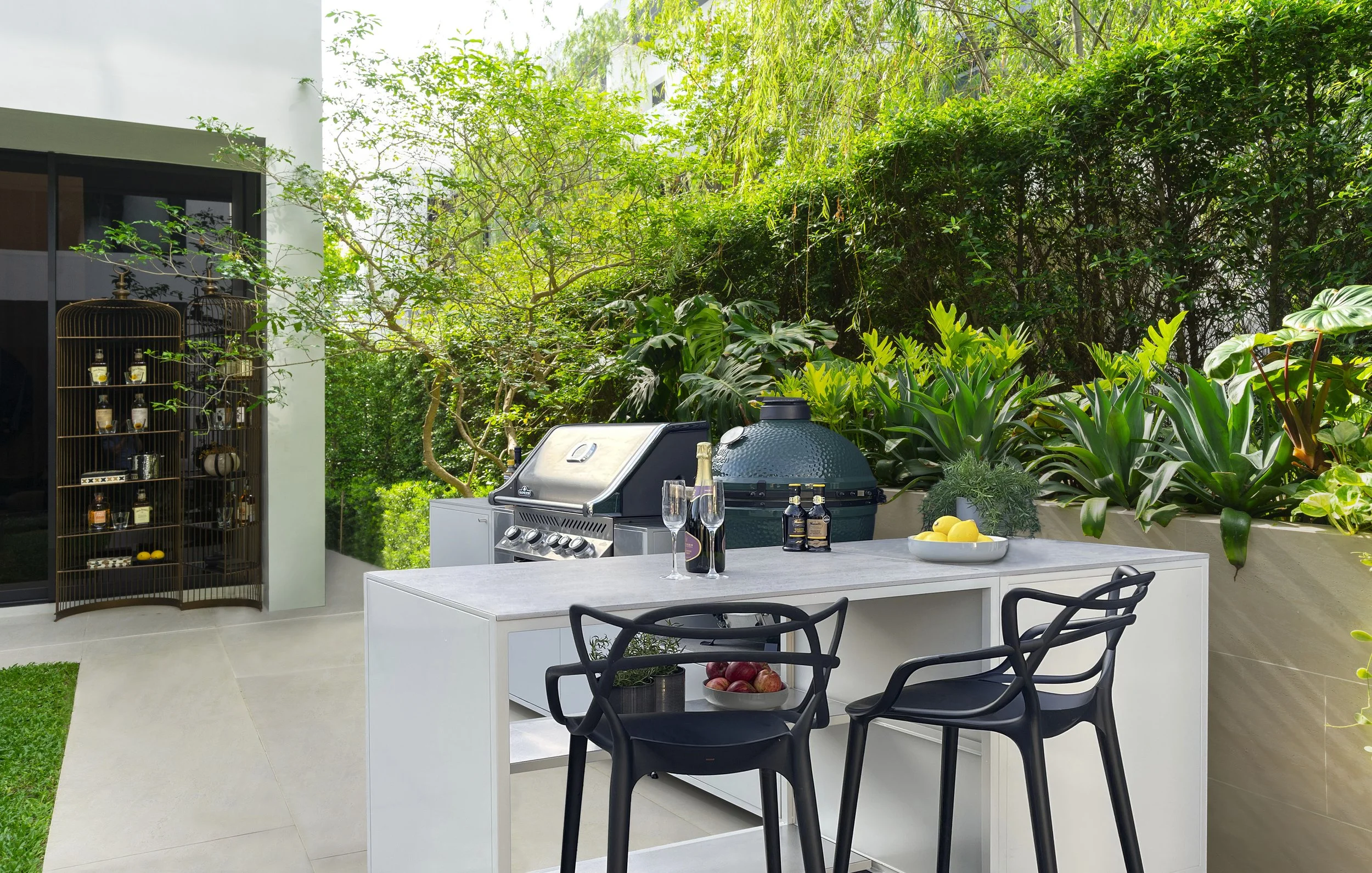 Outdoor patio with a modern grill, a white outdoor table with black chairs, a bowl of lemons, bottles of wine, and two champagne glasses, surrounded by lush green plants and trees.