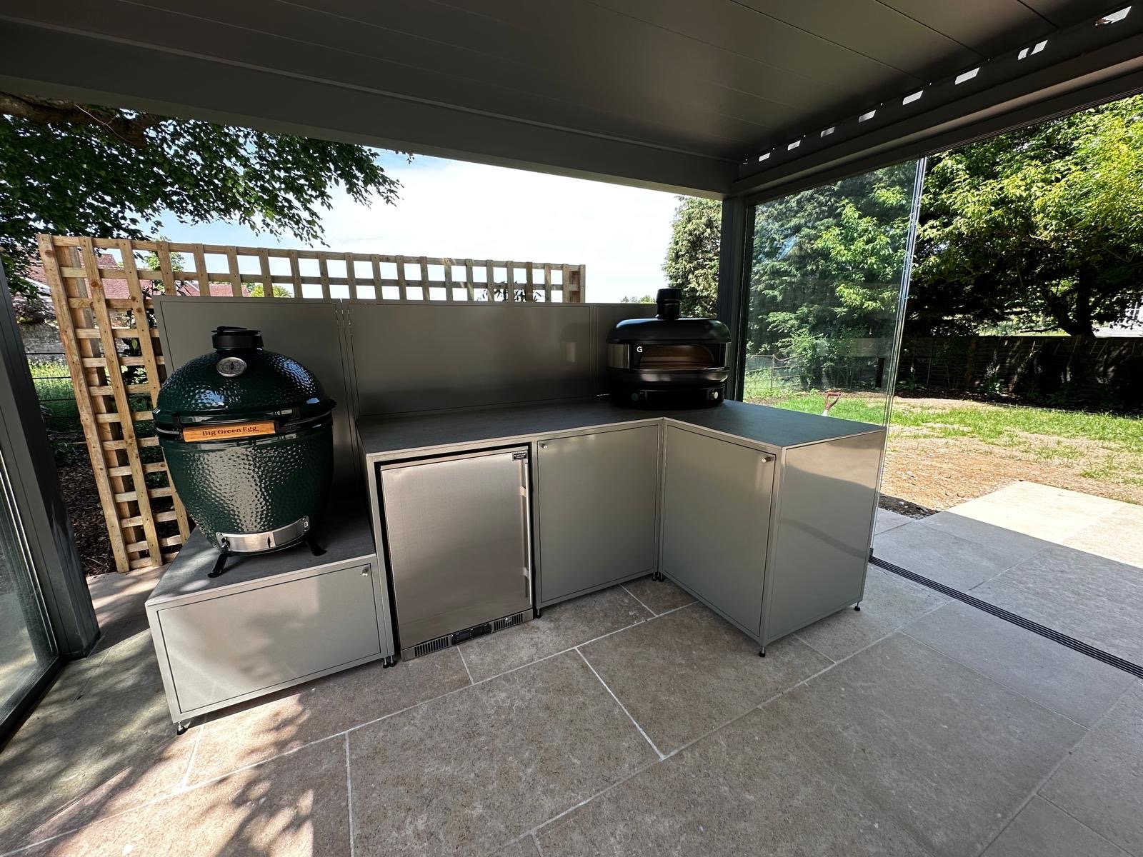 Outdoor kitchen area with a Big Green Egg grill and a black pizza oven on a stainless steel counter, with a backyard visible through glass sliding doors.