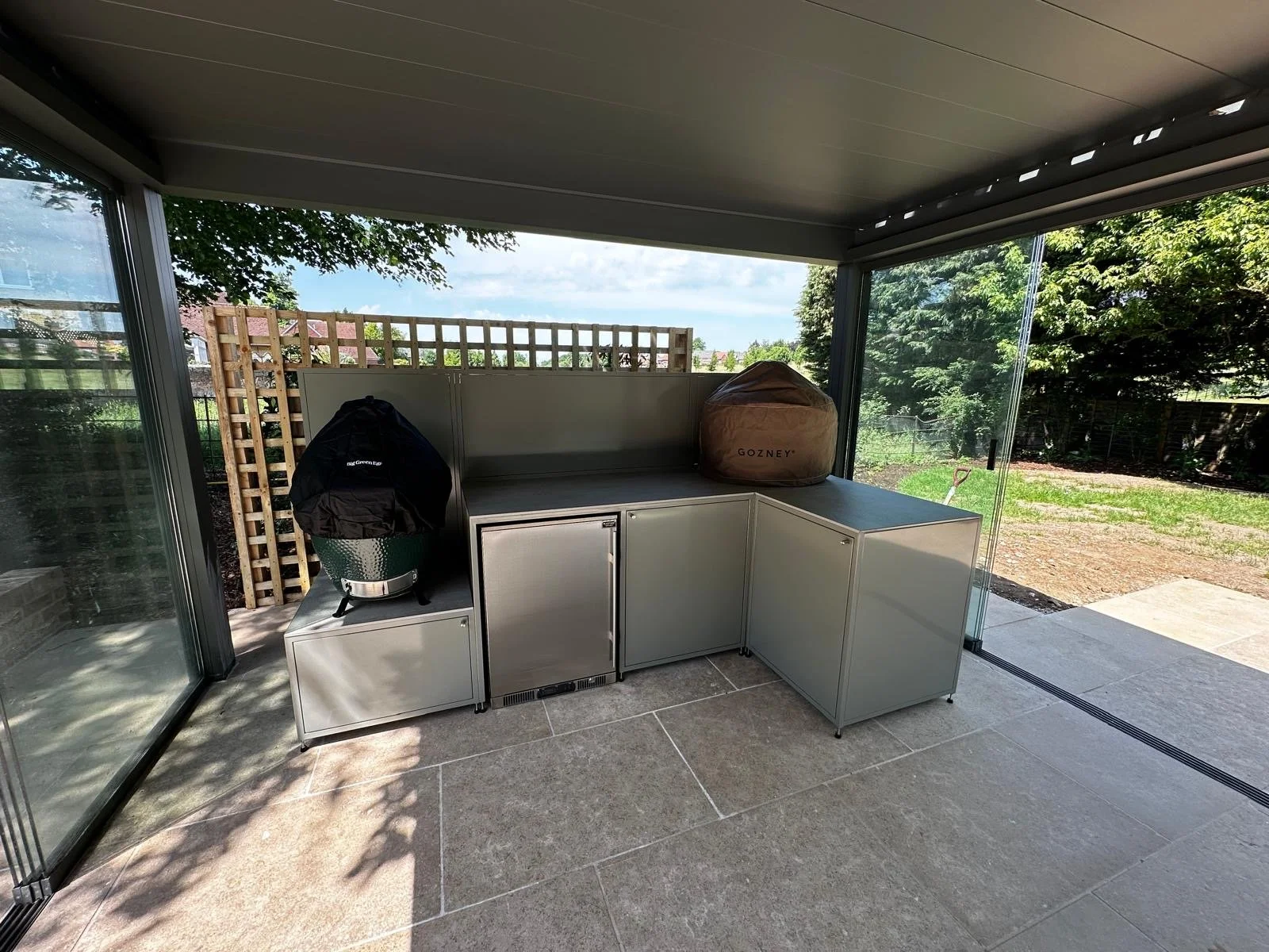 Sunlit outdoor patio with stainless steel storage units, a black grill cover, a brown Gozney pizza oven cover, and greenery outside visible through glass sliding doors.