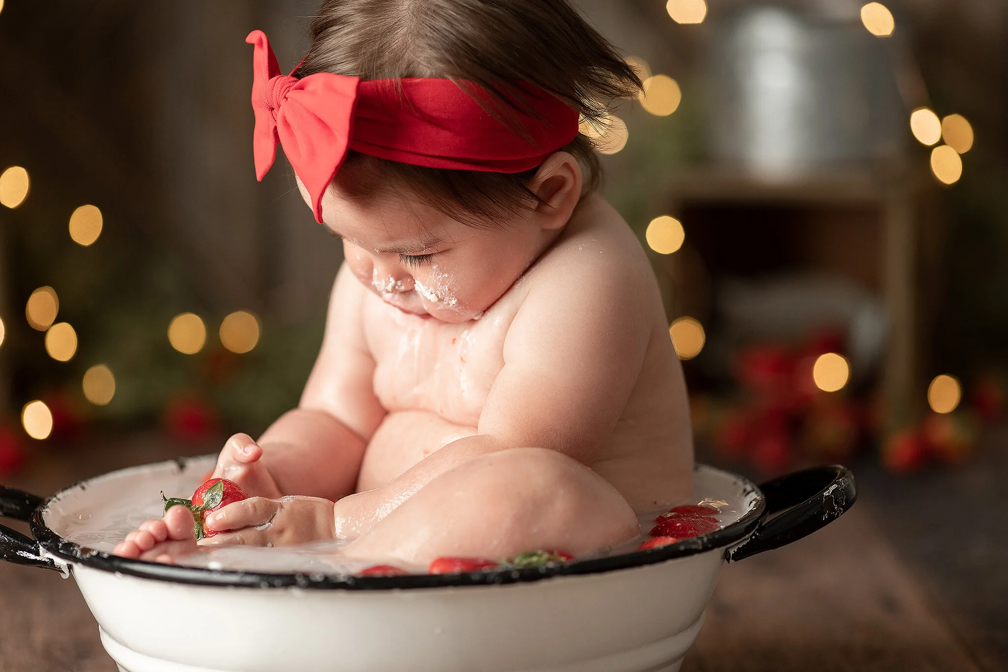 baby girl wearing red bow holding strawberry in milk bath