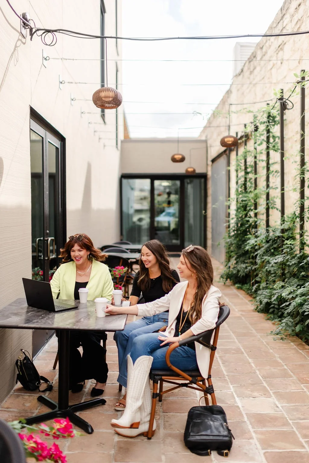 Three women sitting at an outdoor patio table with a laptop, enjoying a coffee break, laughing and chatting, surrounded by greenery and string lights.