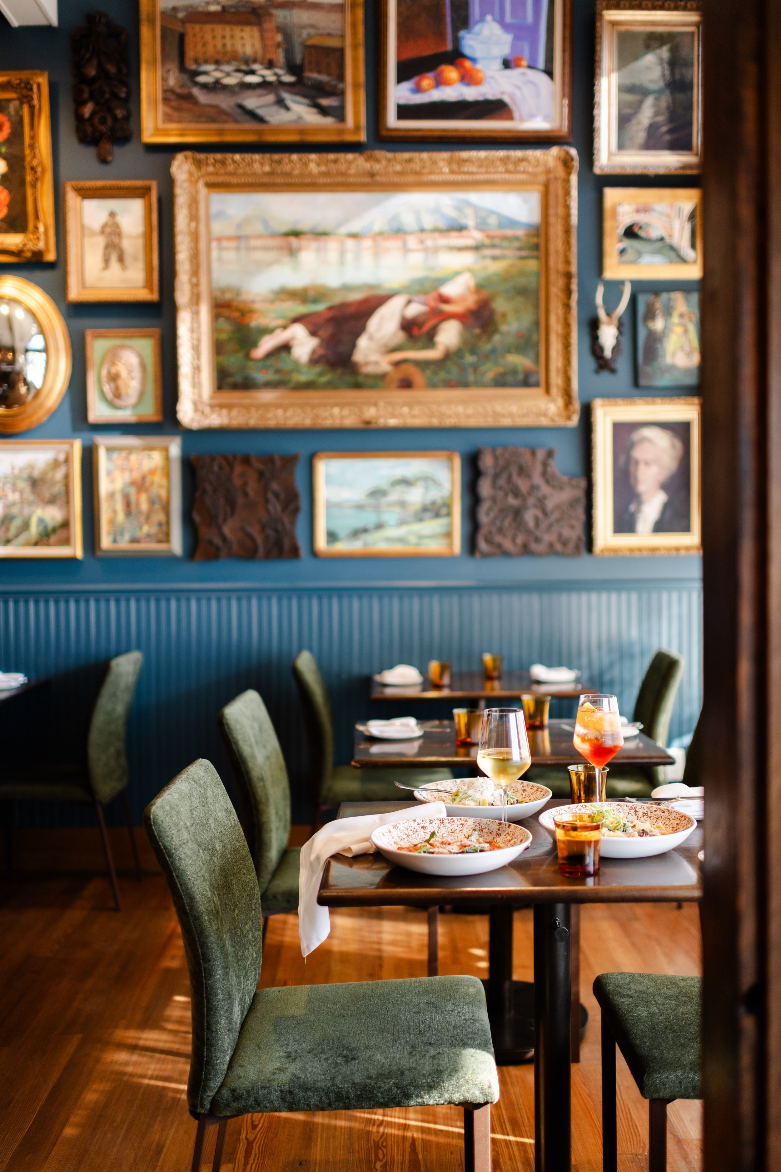 A restaurant dining area with green velvet chairs, wooden tables with plates, glasses, and napkins, and a gallery wall of framed paintings and art pieces.
