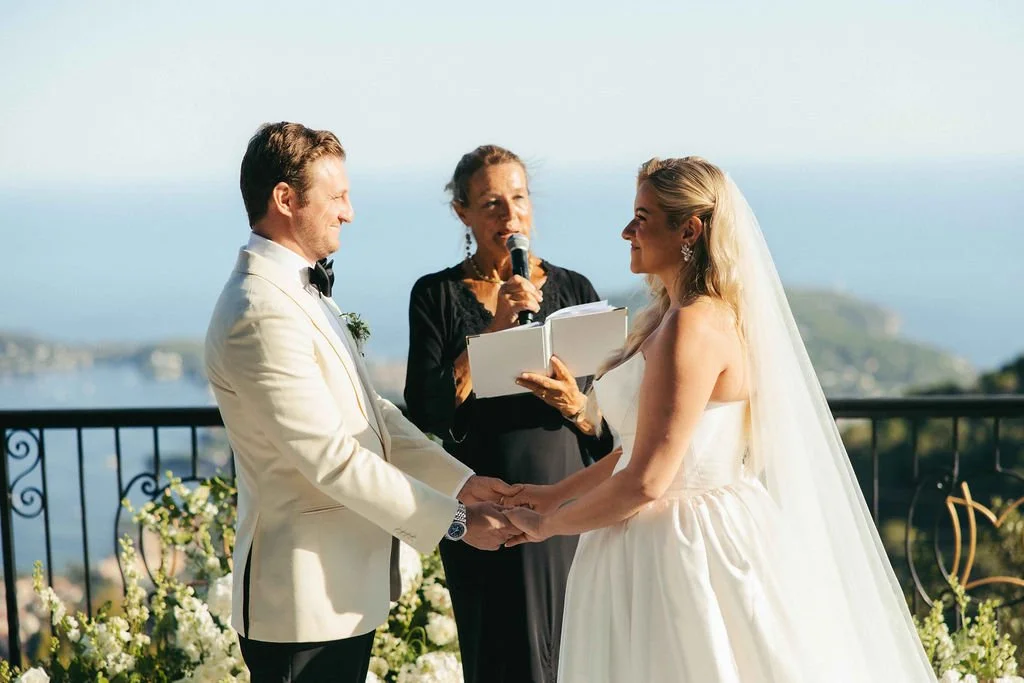 A wedding ceremony with a bride and groom holding hands, standing outdoors on a terrace overlooking water, as an officiant speaks into a microphone.