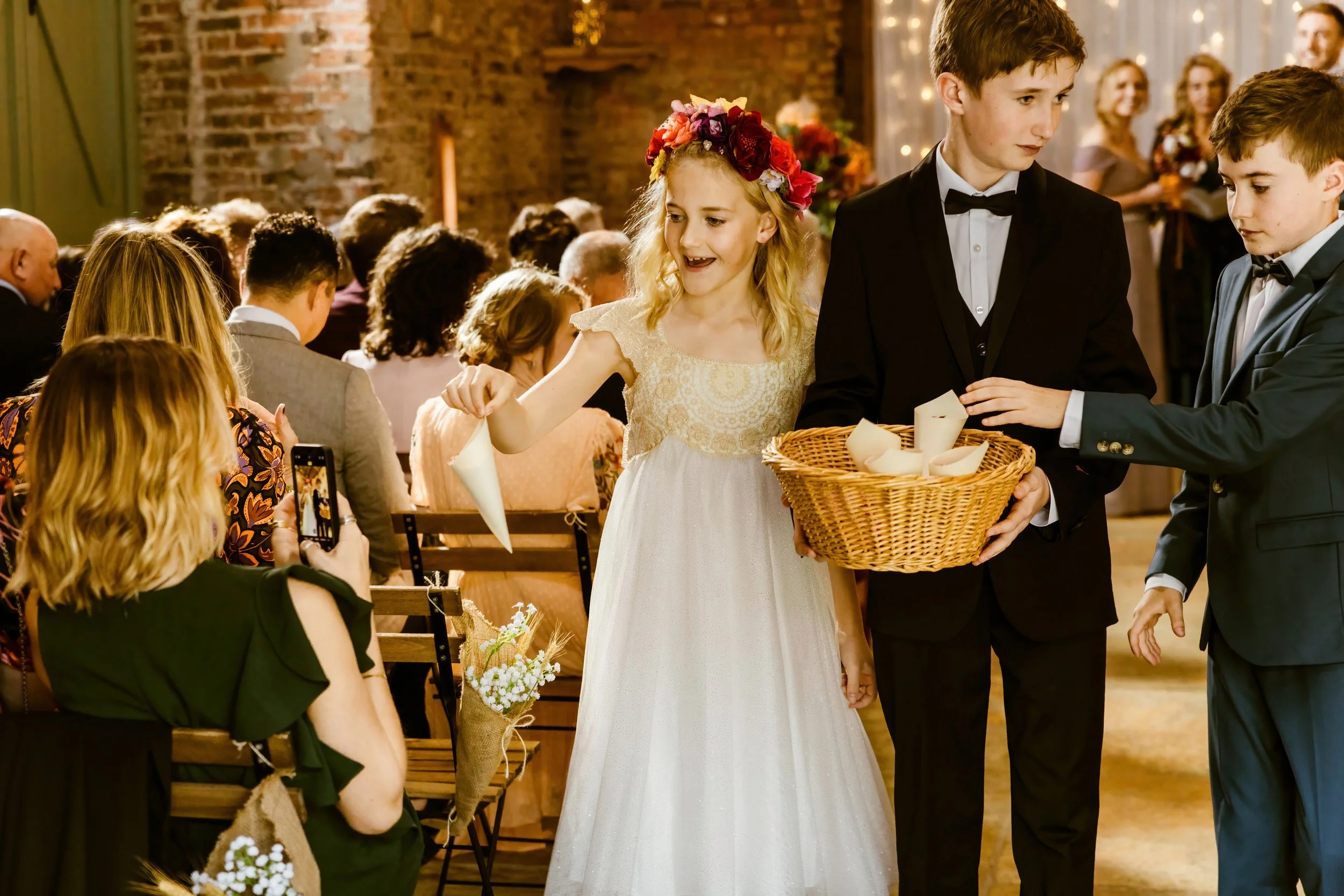 A young girl in a white dress and floral crown is placing a candle in a basket held by a boy in a tuxedo, at a wedding ceremony. Another boy in a tuxedo is assisting him. Guests are seated and watching in the background.