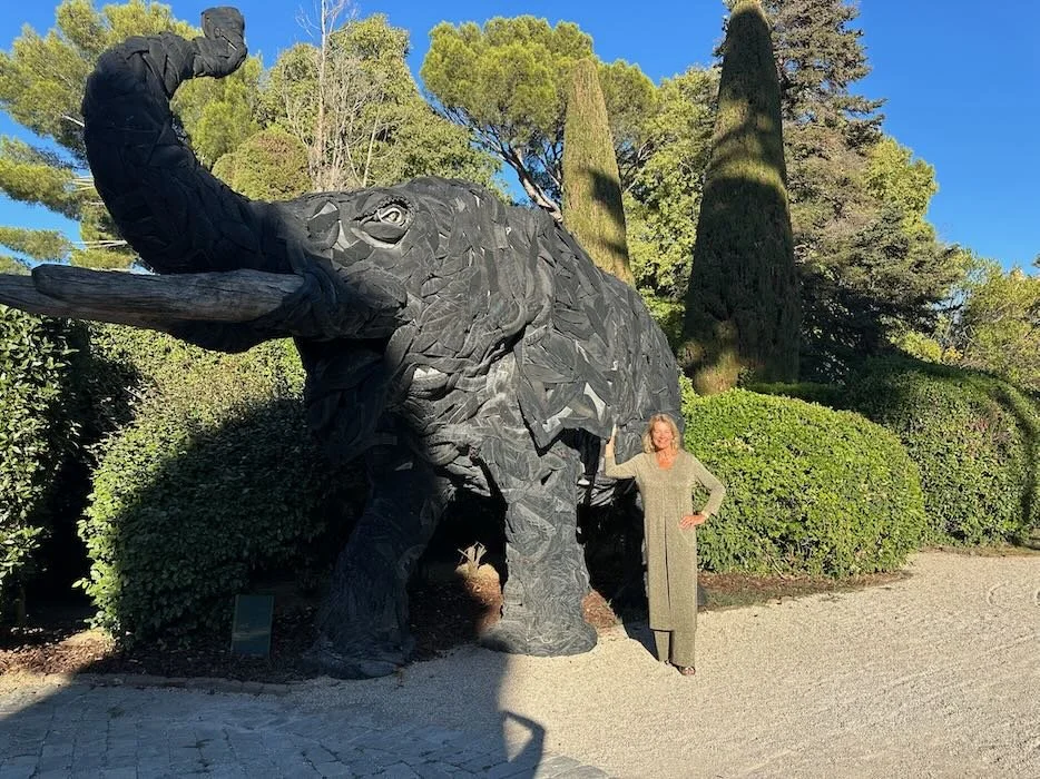 A woman in a green dress standing next to a large wooden statue of an elephant in a park with tall trees and bushes.