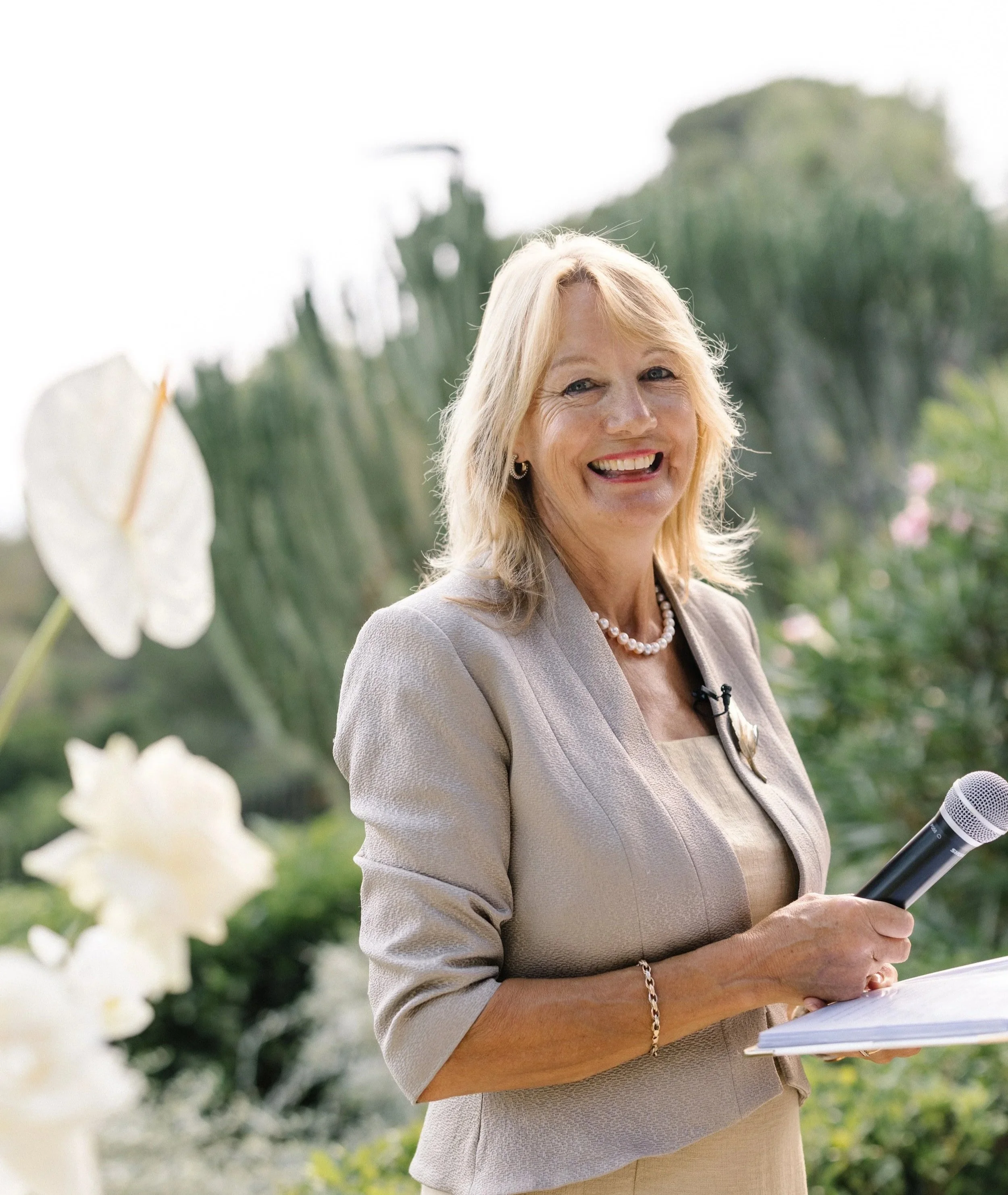 A woman with blonde hair smiling, holding a microphone and a notebook, standing outdoors with green foliage and white flowers in the background.