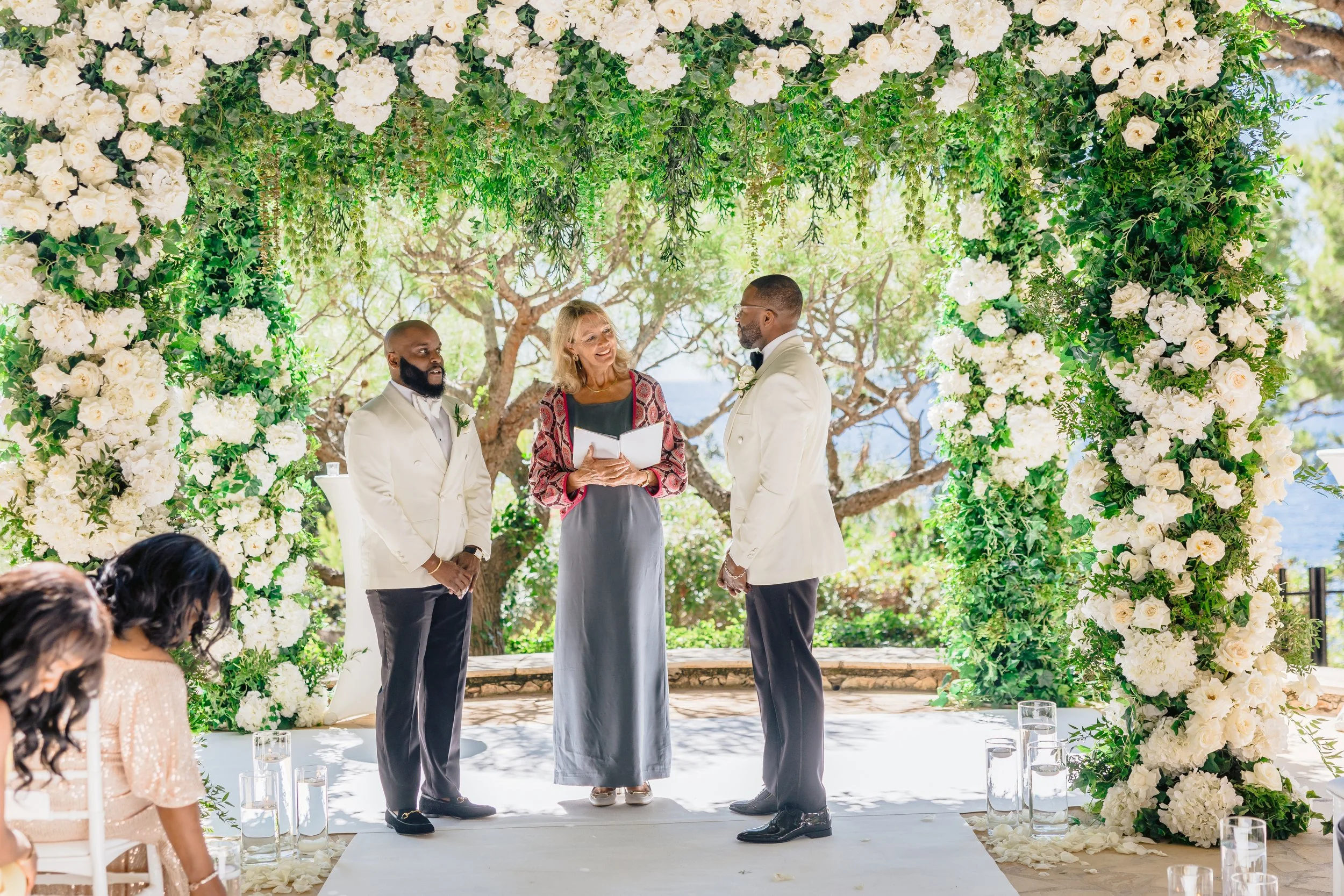 A wedding ceremony taking place outdoors, with a couple exchanging vows under an arch decorated with white flowers and greenery. An officiant is standing between them, holding a book. Guests are seated nearby, and the background features trees and a clear sky.