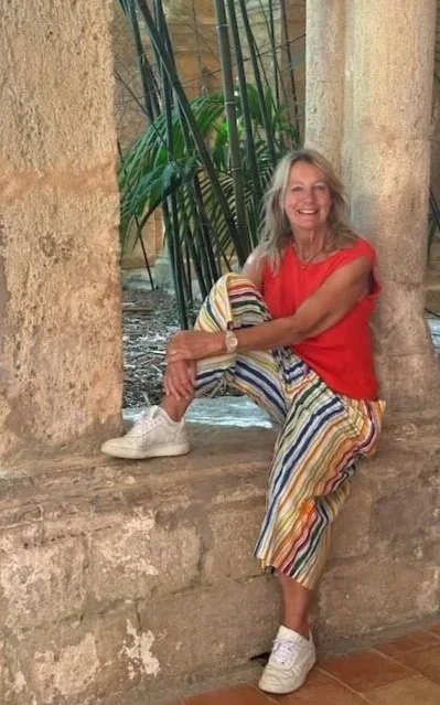 A smiling middle-aged woman with long gray hair sitting on a stone ledge between two large stone columns, wearing a bright red sleeveless top, colorful striped pants, and white sneakers. She is outdoors with green plants behind her.