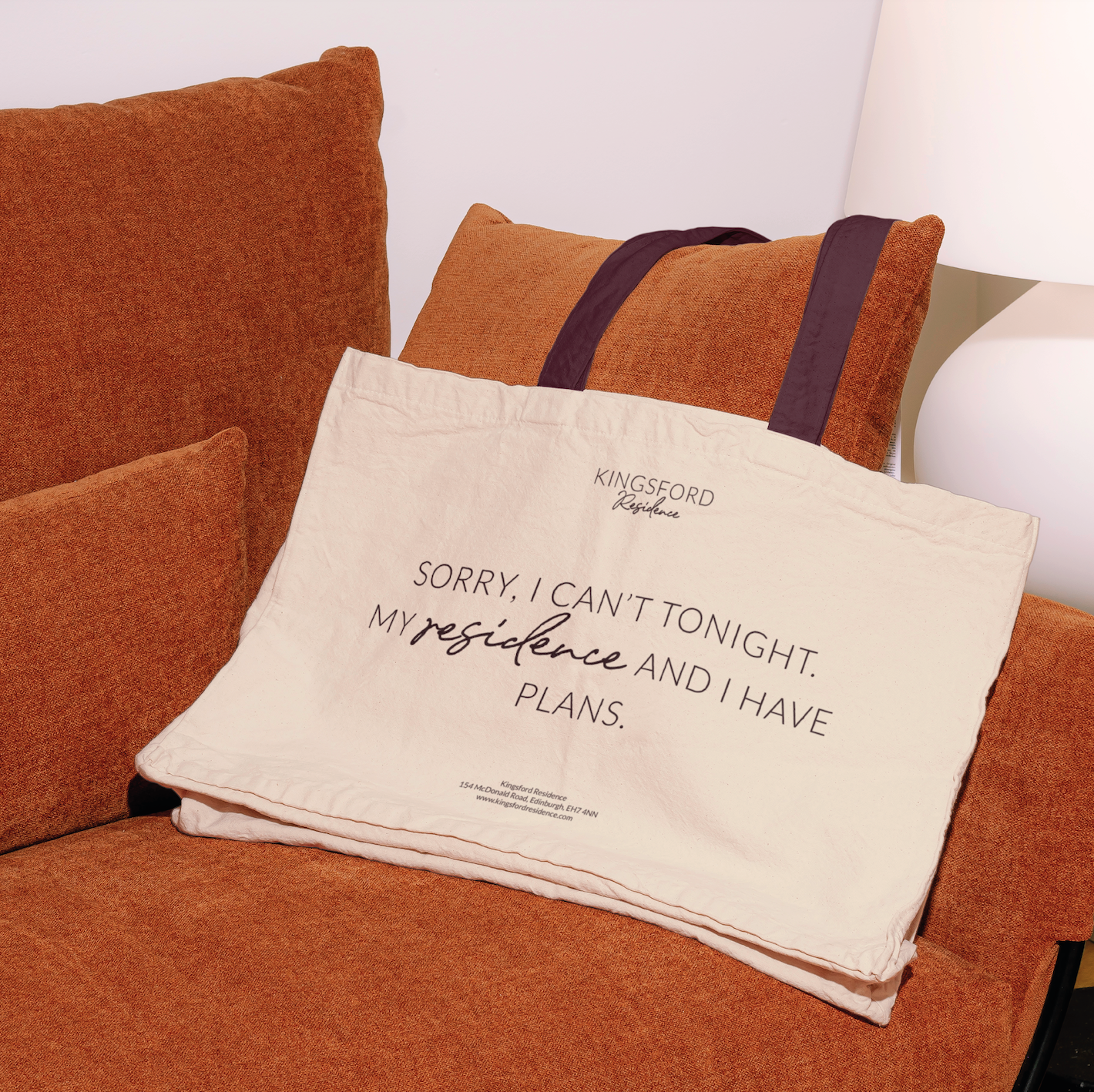 A beige fabric tote bag with black handles resting on an orange upholstered sofa. The bag has text that reads, "KINGSFORD Residence. Sorry, I can't tonight. My residence and I have plans." next to a small printed address.