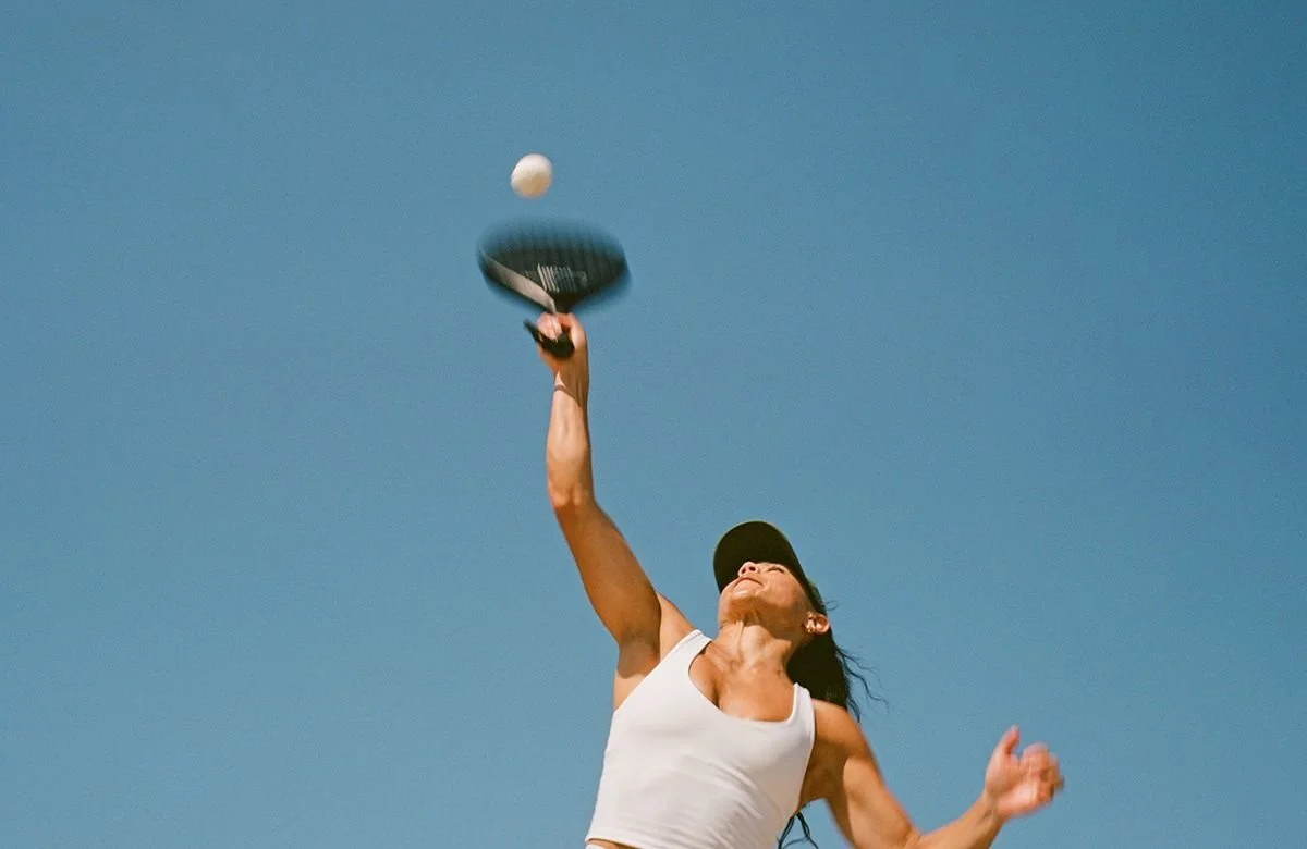 A woman wearing a white tank top and black cap playing tennis outdoors, reaching up to hit a tennis ball with a racket against a clear blue sky.