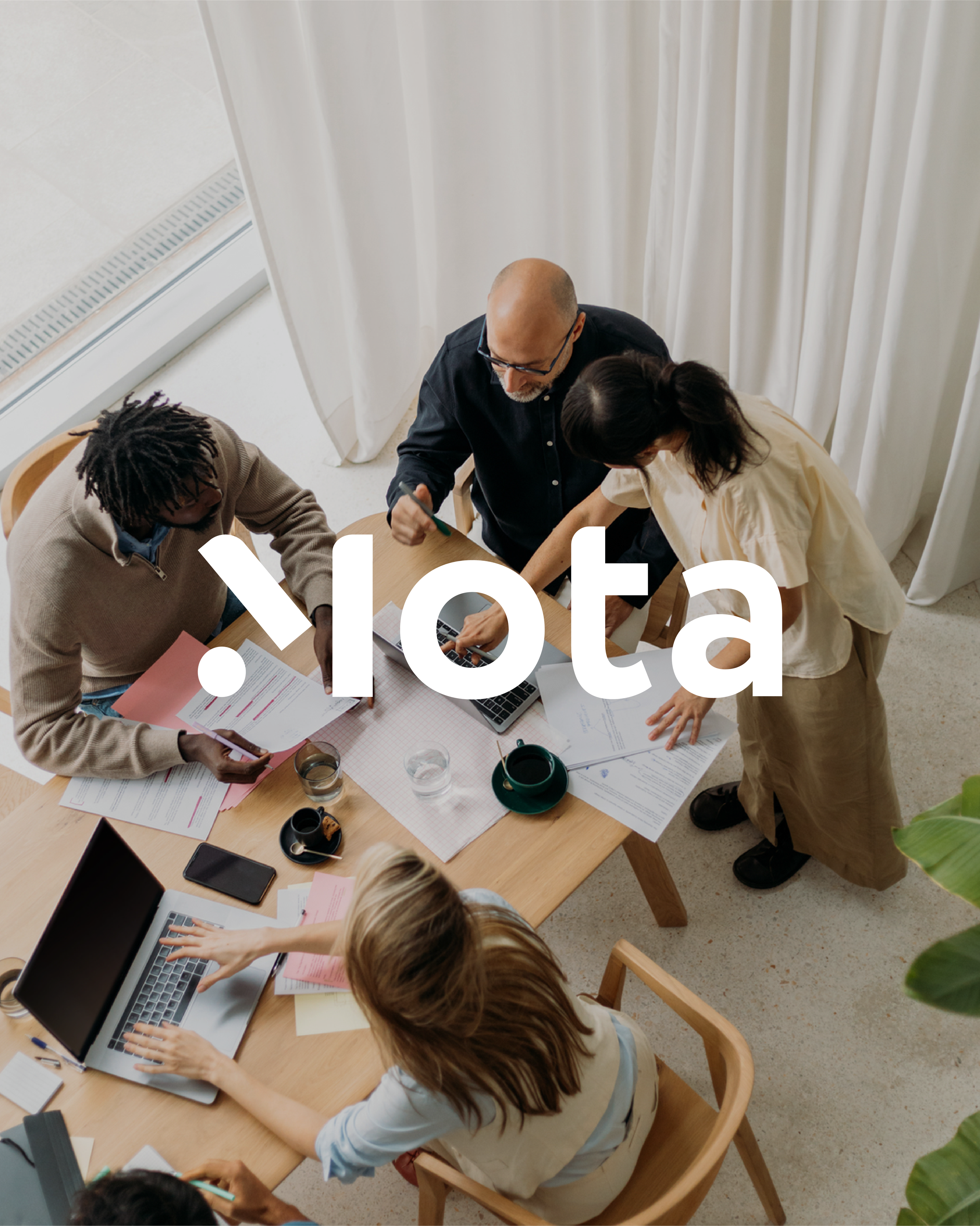 A group of six people gathered around a wooden table working on documents and laptops in a well-lit room with white curtains and a tiled floor.
