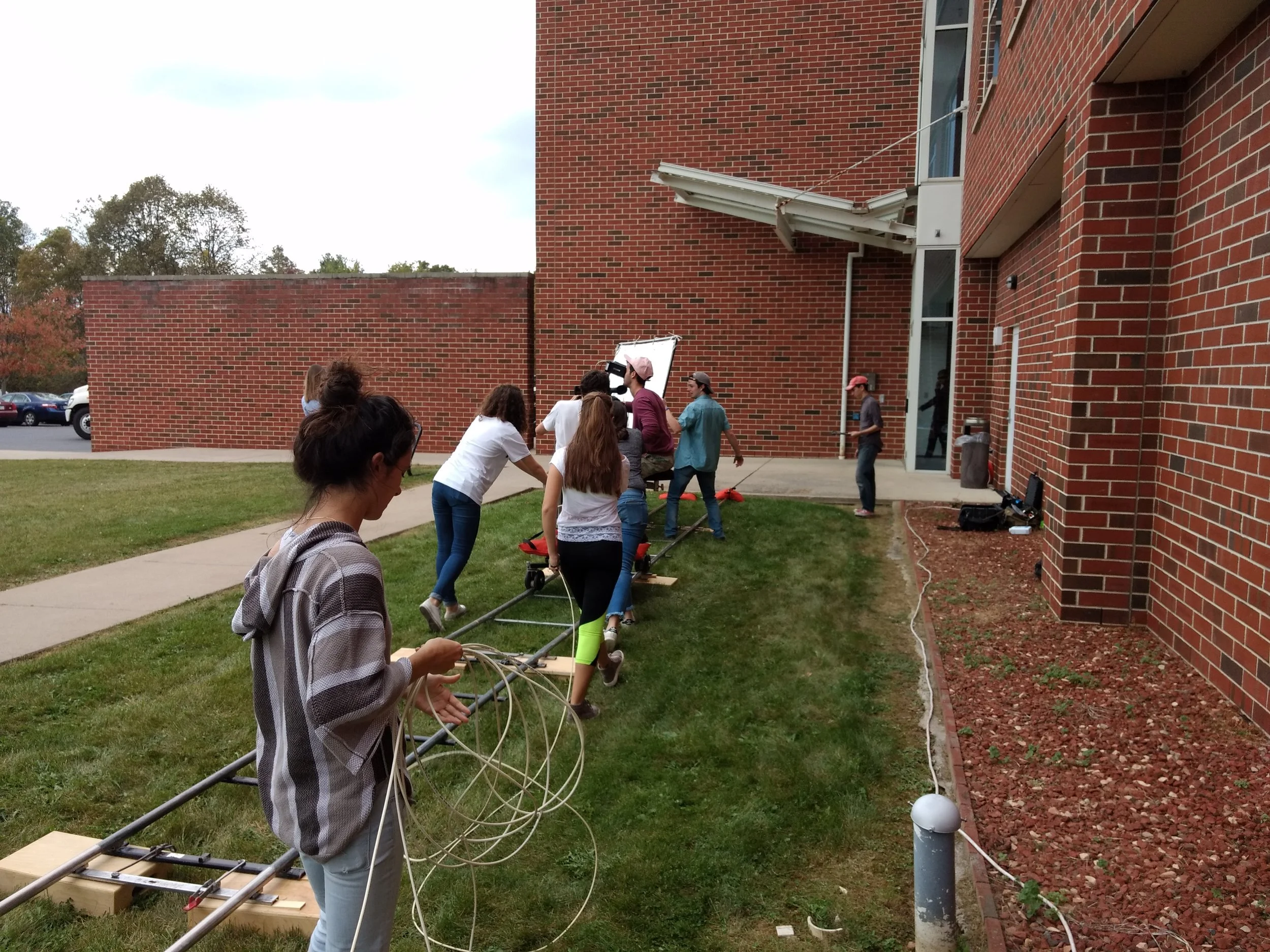 Group of people working outdoors on a film or television production set, with some pulling a long cable or rope along the grass outside a brick building.