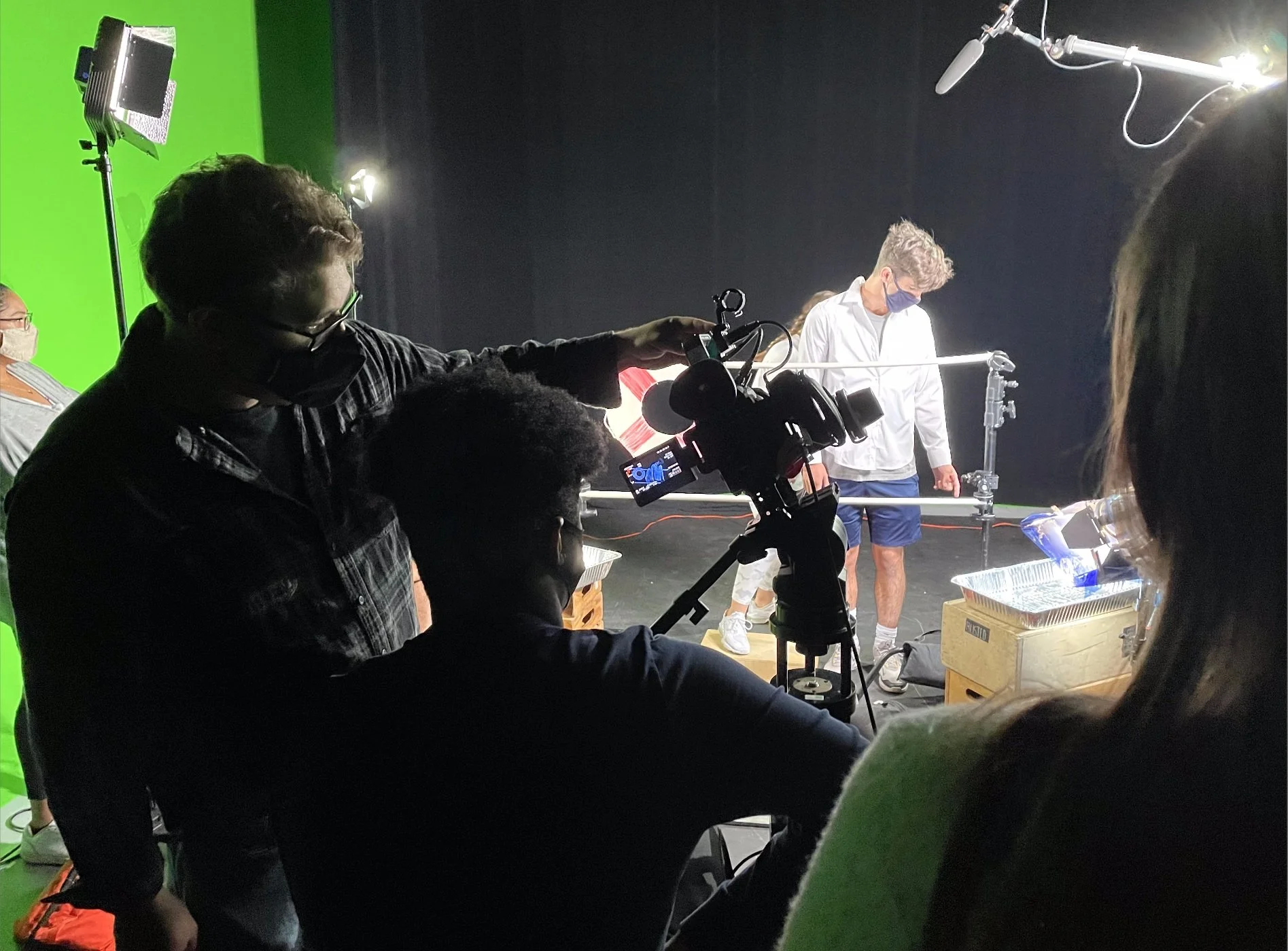 Film crew recording scene with two people, one in white shirt and shorts and another in red shirt, surrounded by equipment including cameras, lights, and a green screen.