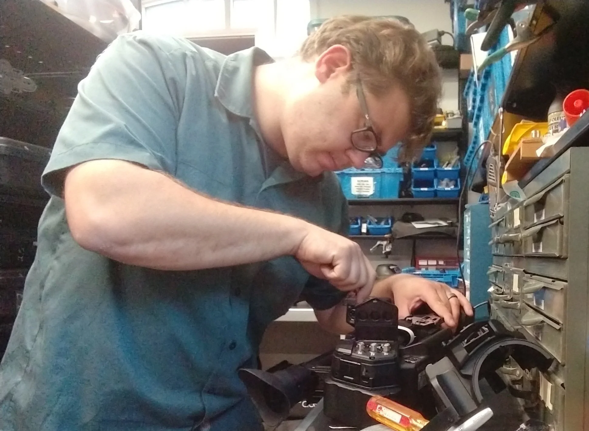 A man wearing glasses and a gray shirt working with electronic equipment and tools in a workshop or repair shop.