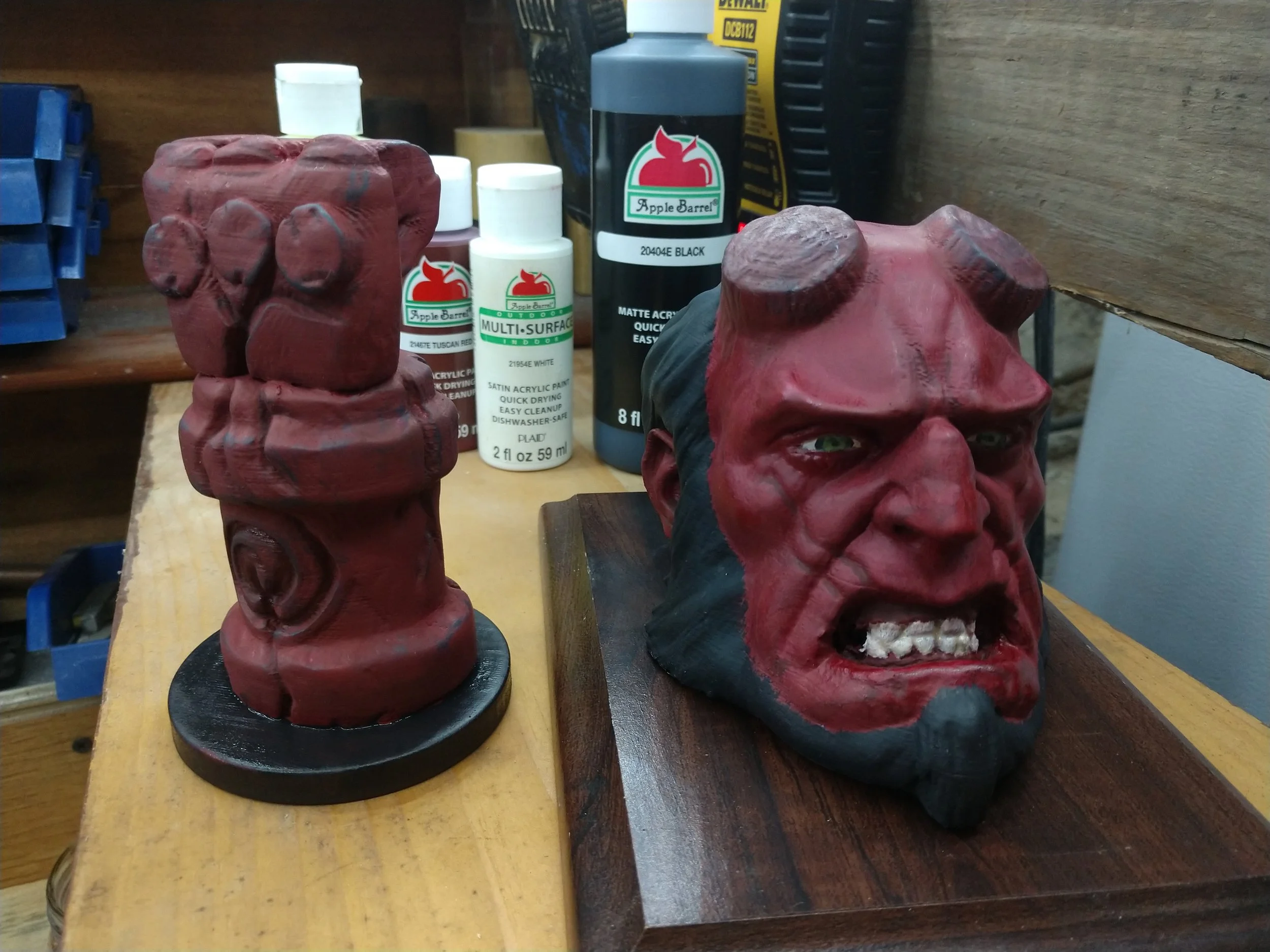 A sculpture of the Hulk's head with an angry expression, clenched teeth, and a black beard, mounted on a wooden base, with a red painted body chiseled into a fist next to it, surrounded by paint bottles in a workshop.
