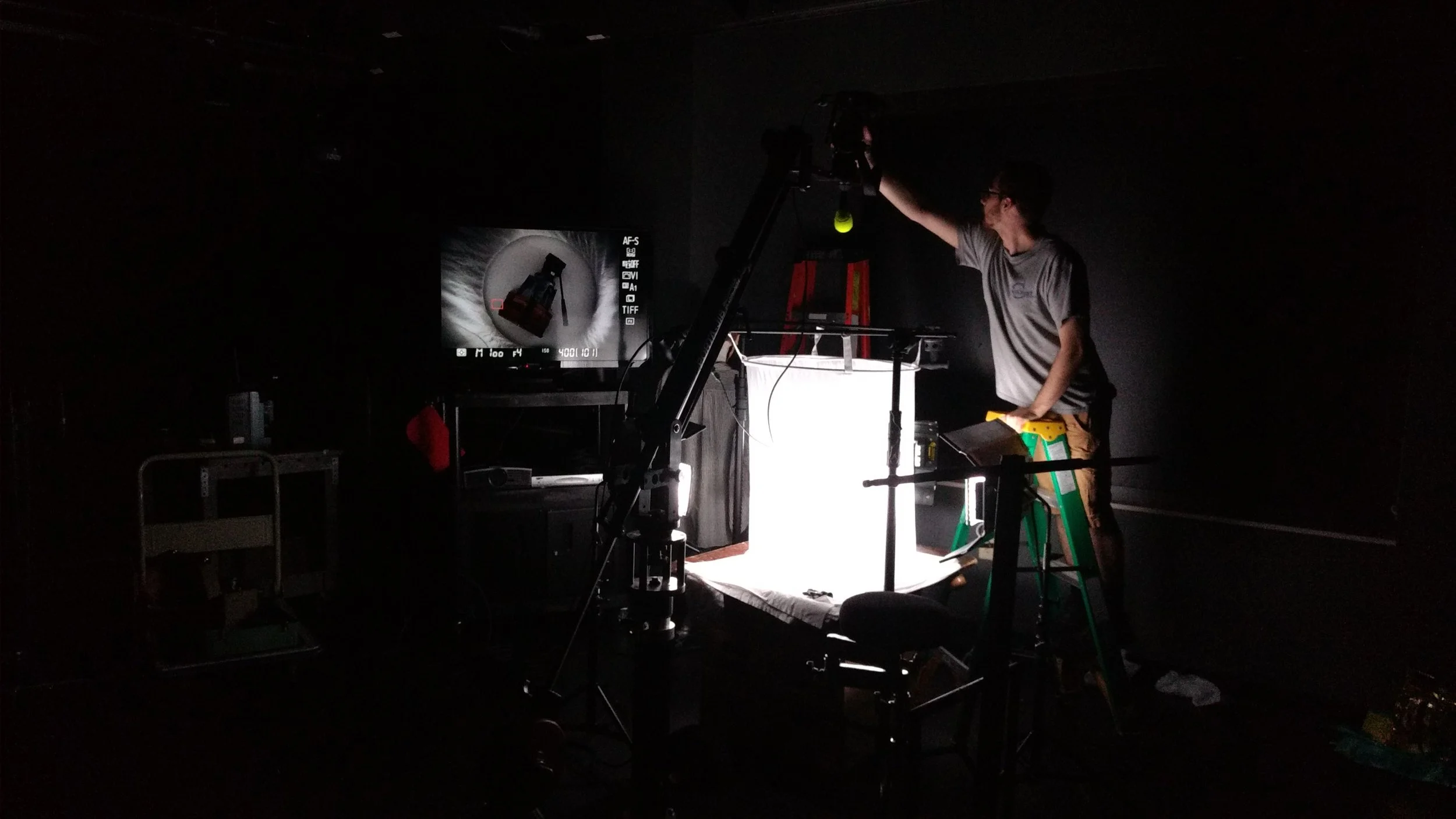 A person working in a dark room, lit by a bright light, surrounded by photography equipment including a camera, tripod, and ladder, with a monitor displaying a camera view.