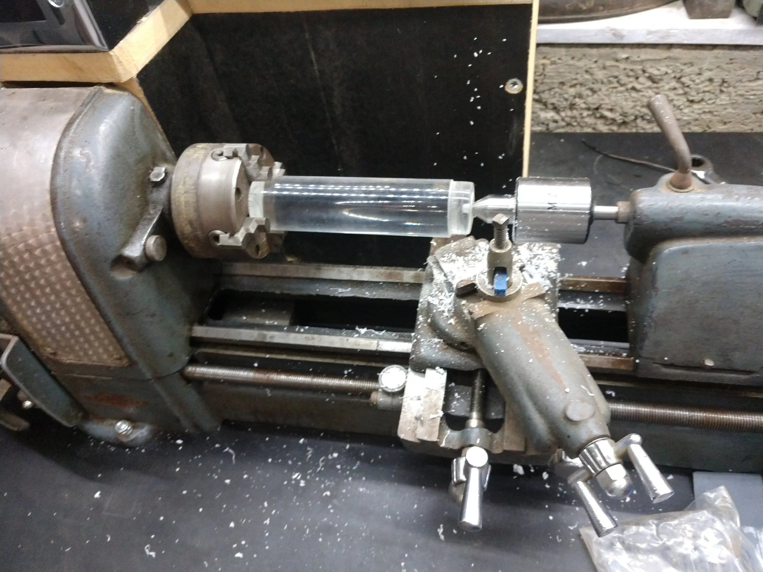 A metal lathe machine with a cylindrical glass tube mounted on it, surrounded by metal shavings and cluttered workshop surface.