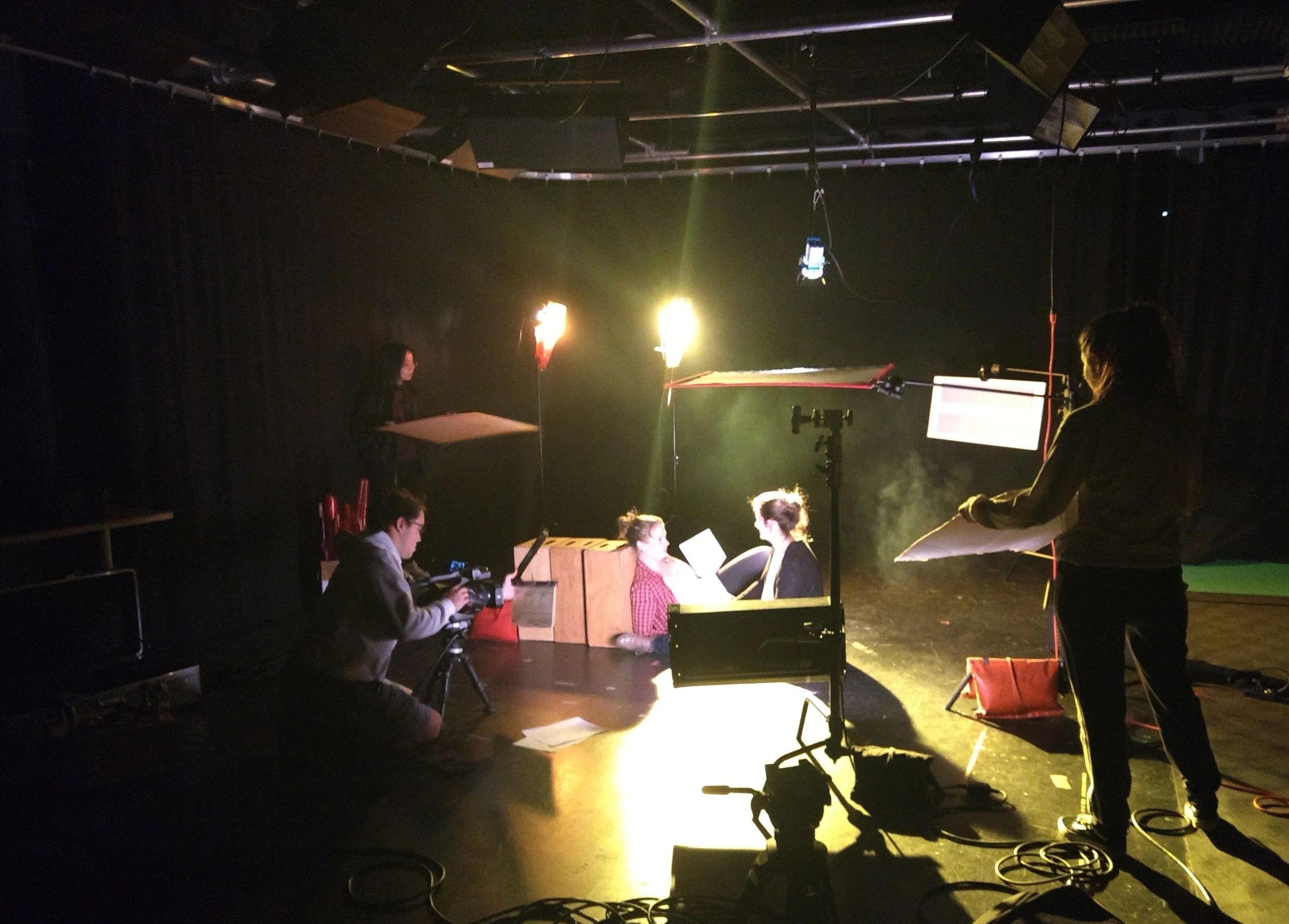 Lighting and camera crew filming in a dark studio with black curtains, surrounding by professional filming equipment and crew members.