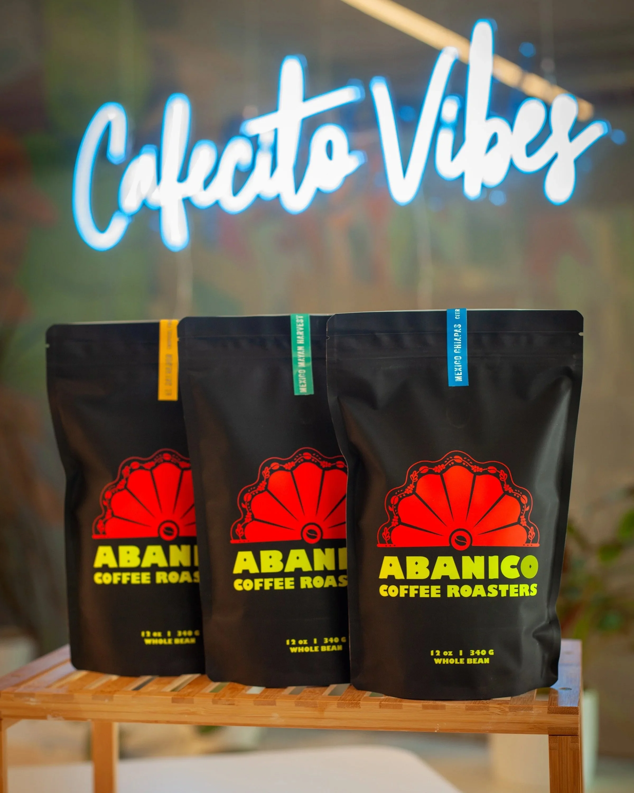 Three bags of Abancó coffee roasters on a wooden shelf with a blurred background and a neon sign that says 'cactus Vibes'.
