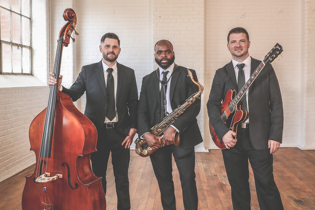 Three men in suits holding musical instruments, standing indoors on a wooden floor in front of a brick wall with large windows.